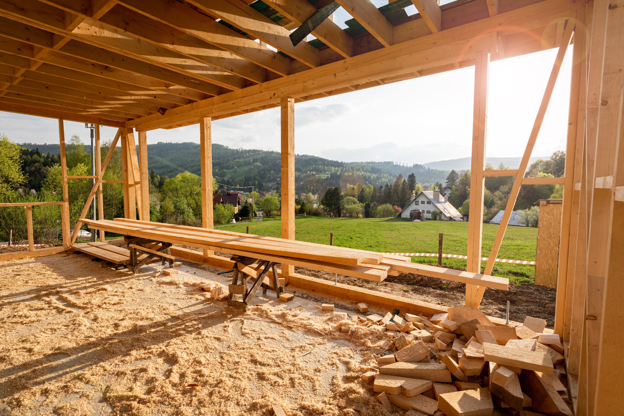 Wnętrze w trakcie budowy domu z drewnianą konstrukcją szkieletową, z widokiem na krajobraz z domami w tle, oświetlone popołudniowym słońcem.