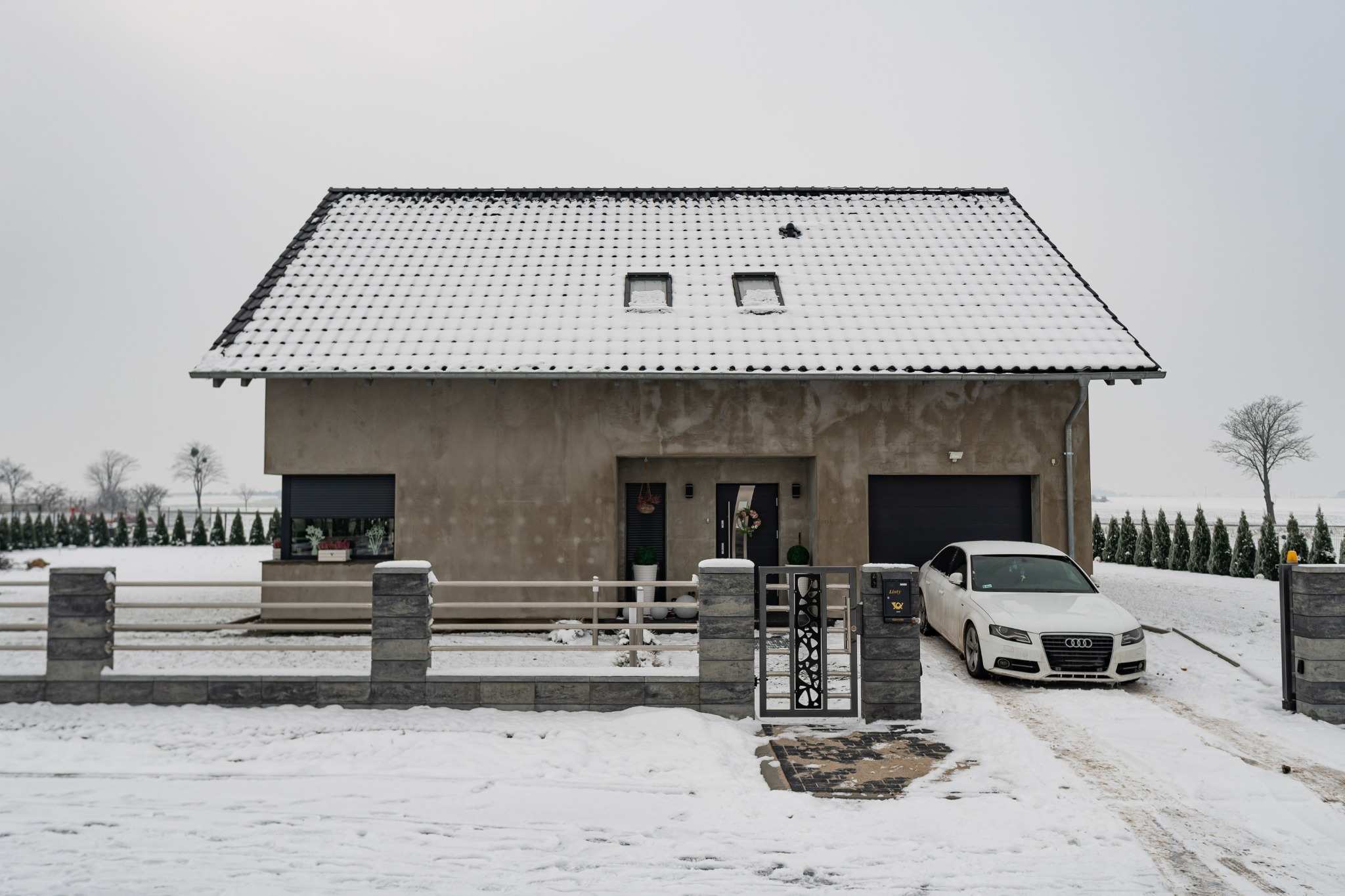 Surowy, nowoczesny dom z płaskim dachem pokrytym śniegiem, z szarym ogrodzeniem i białym samochodem na ośnieżonym podjeździe w zimowy dzień.