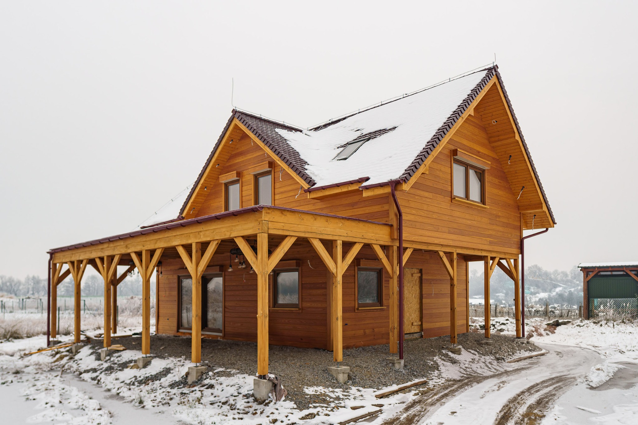Zimowy krajobraz z drewnianym domem na podwyższeniu, z zadaszonym tarasem, częściowo pokrytym śniegiem.