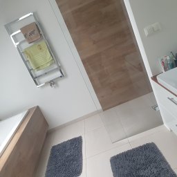 Czarodziejka - Nowoczesna łazienka z wanną, grzejnikiem drabinkowym z ręcznikami, szklaną kabiną prysznicową, umywalką z szafką i dwoma szarymi dywanikami na białych płytkach.