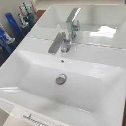 Czarodziejka - Biała, prostokątna umywalka z chromowaną baterią, odbijająca się w lustrze, z akcesoriami łazienkowymi w tle i mydłem w mydelniczce po prawej stronie.