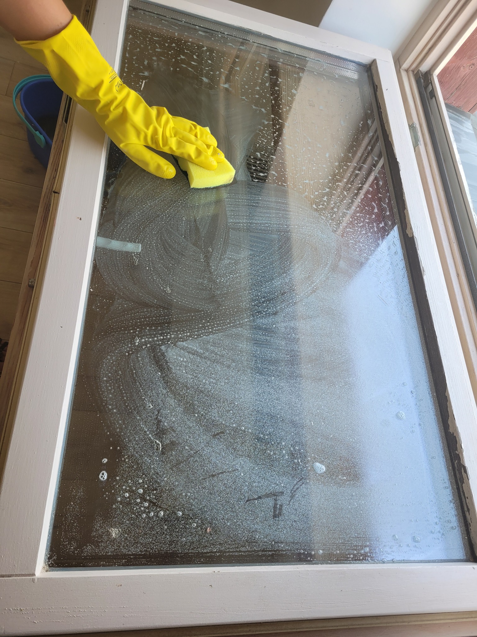 Żółta rękawica z gąbką myje okno z białymi piankowymi smugami, widoczny fragment niebieskiego wiadra.