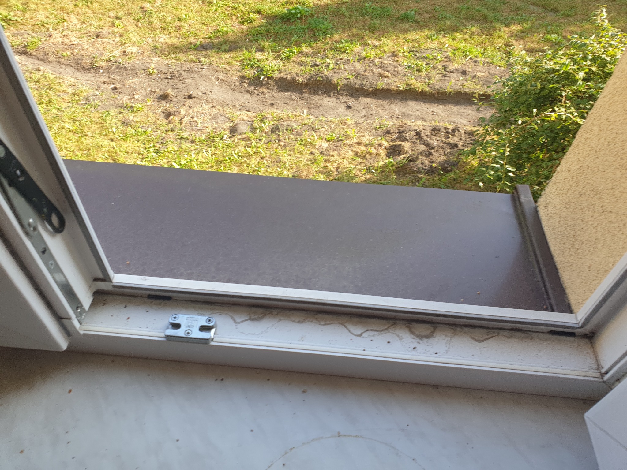 Widok z bliska na brudną, białą ramę okienną z widocznymi śladami po zaciekach i kurzu, z zielenią i ziemią widoczną za oknem.