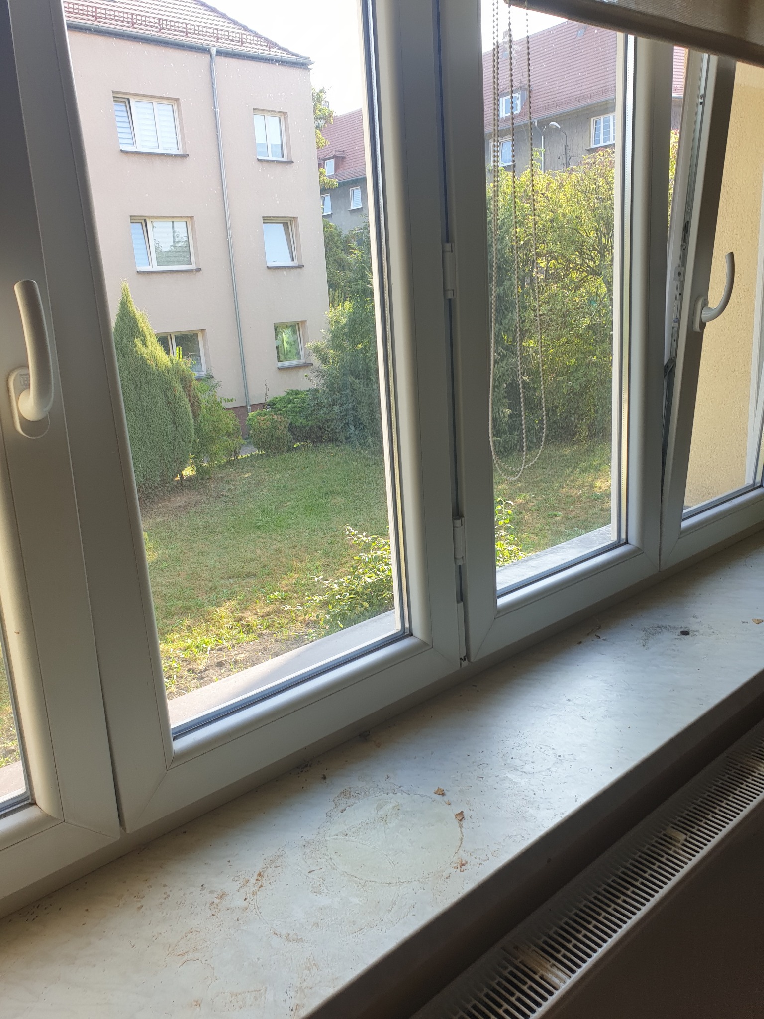 Białe, trzyskrzydłowe okno PCV z widokiem na zielony ogród i budynki. Parapet okienny z widocznymi zabrudzeniami i plamami, poniżej grzejnik.