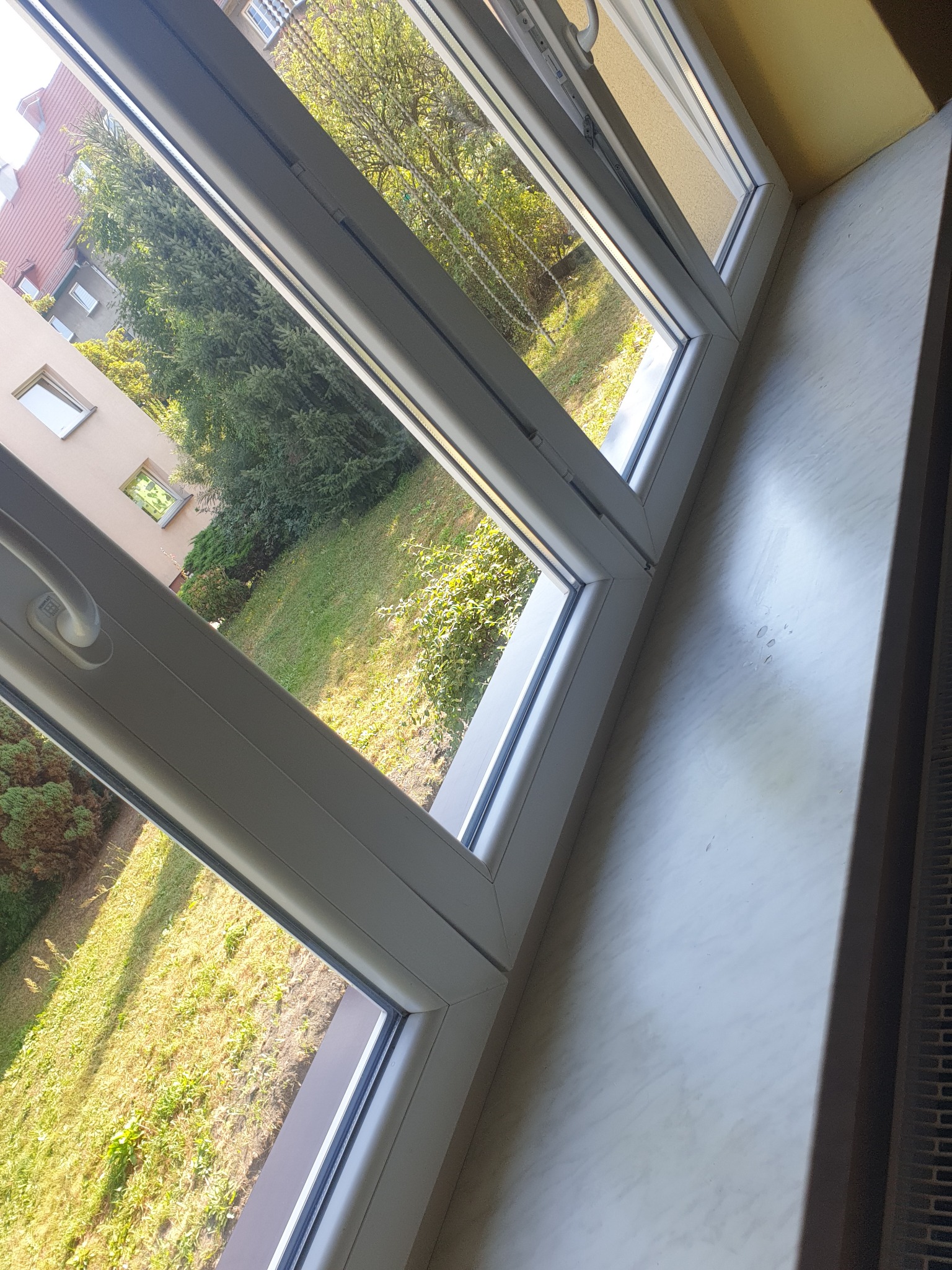 Otworzone okno PCV z widokiem na zielony ogród z trawnikiem i drzewami, widoczne ślady po kroplach na parapecie.