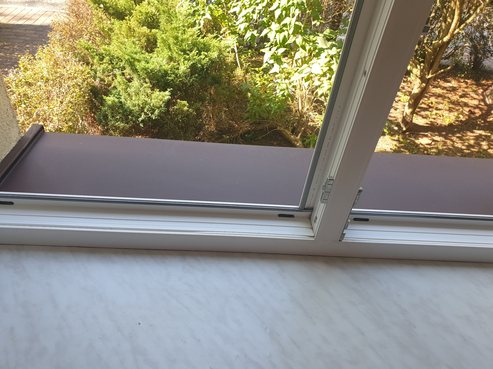 Otwarty fragment okna z widokiem na zieleń na zewnątrz, widoczny parapet wewnętrzny z wzorem marmurkowym.