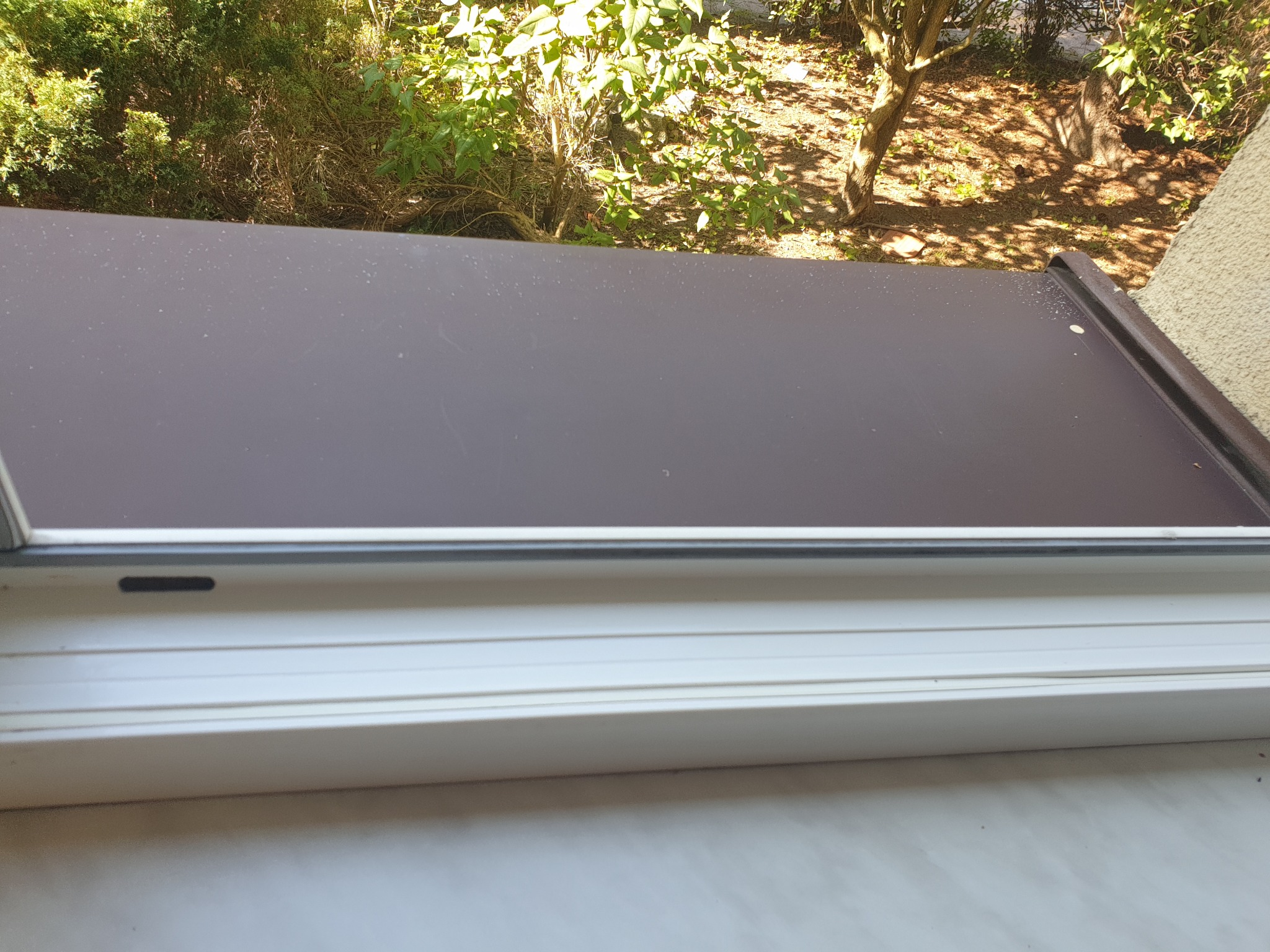 Zabrudzona, brązowa zewnętrzna roleta okienna przed umyciem, widok na zieloną roślinność za oknem.