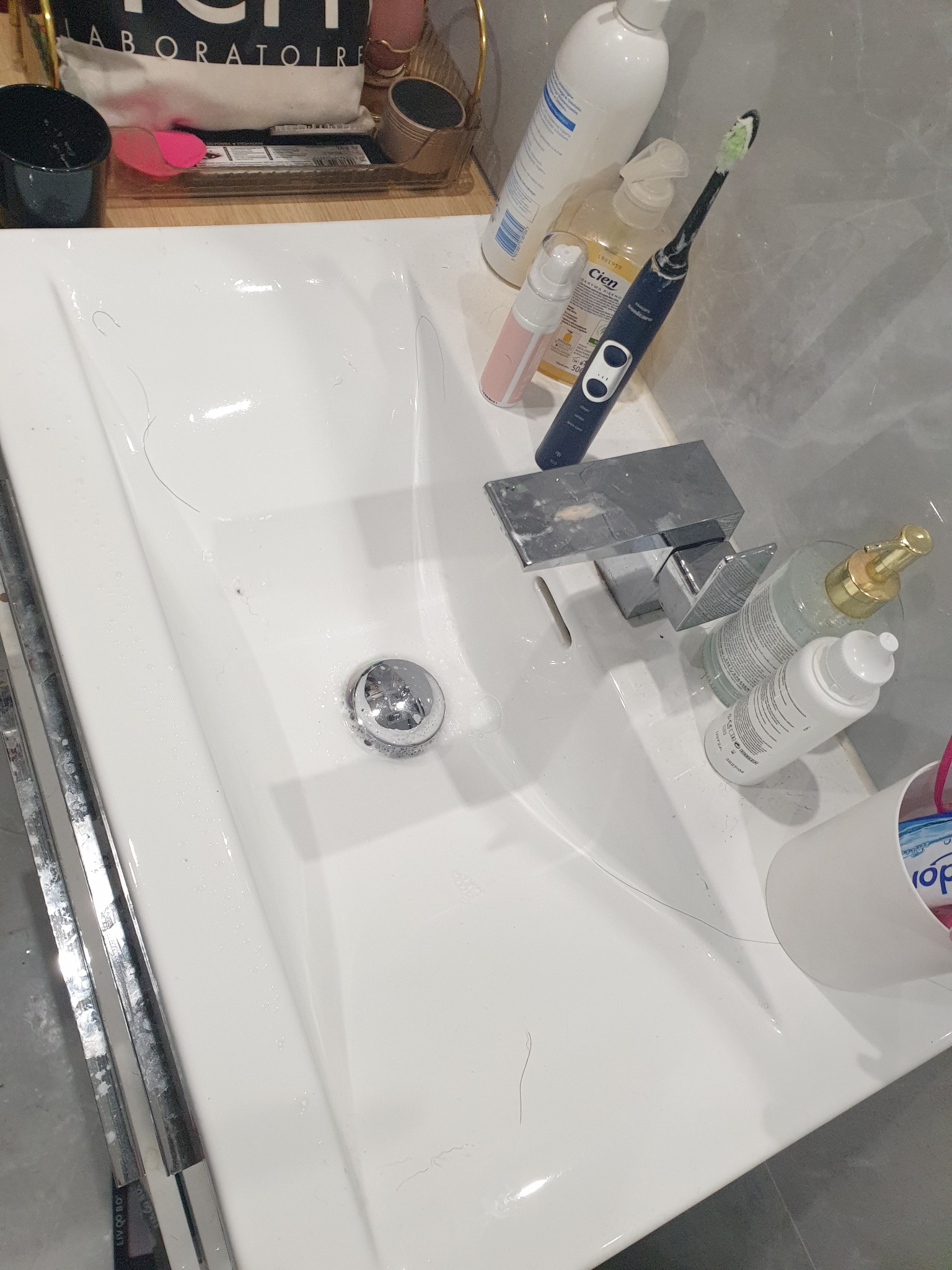 Biała umywalka z chromowaną baterią, na której leżą butelki kosmetyków, elektryczna szczoteczka do zębów i widoczne pojedyncze włosy. Umywalka częściowo mokra.