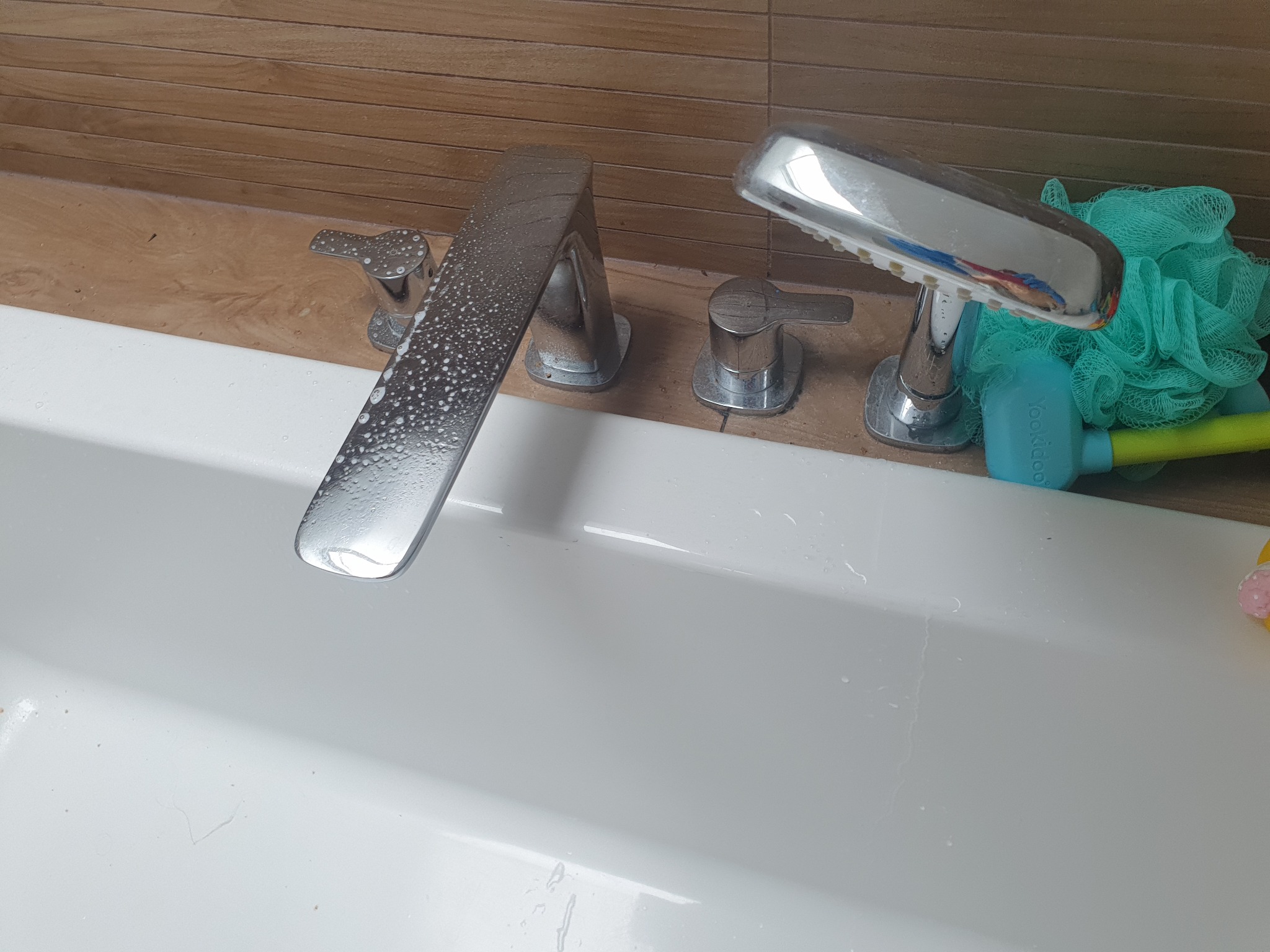 Bateria wannowa z chromowanym wykończeniem pokryta kroplami wody, obok leży zielona myjka i niebieska zabawka do kąpieli, widoczne ślady czyszczenia na białej powierzchni wanny.