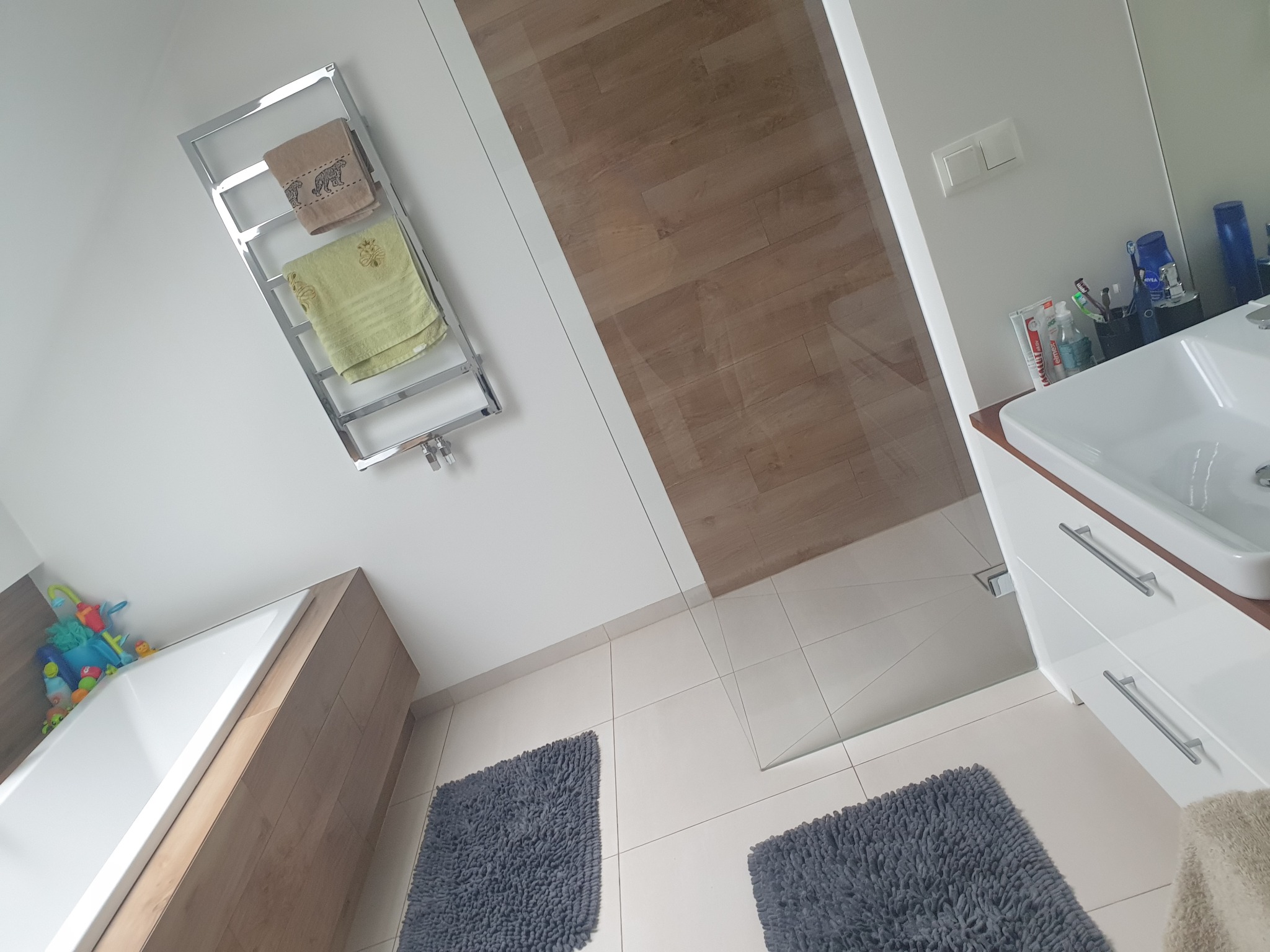 Nowoczesna łazienka z wanną, grzejnikiem drabinkowym z ręcznikami, szklaną kabiną prysznicową, umywalką z szafką i dwoma szarymi dywanikami na białych płytkach.