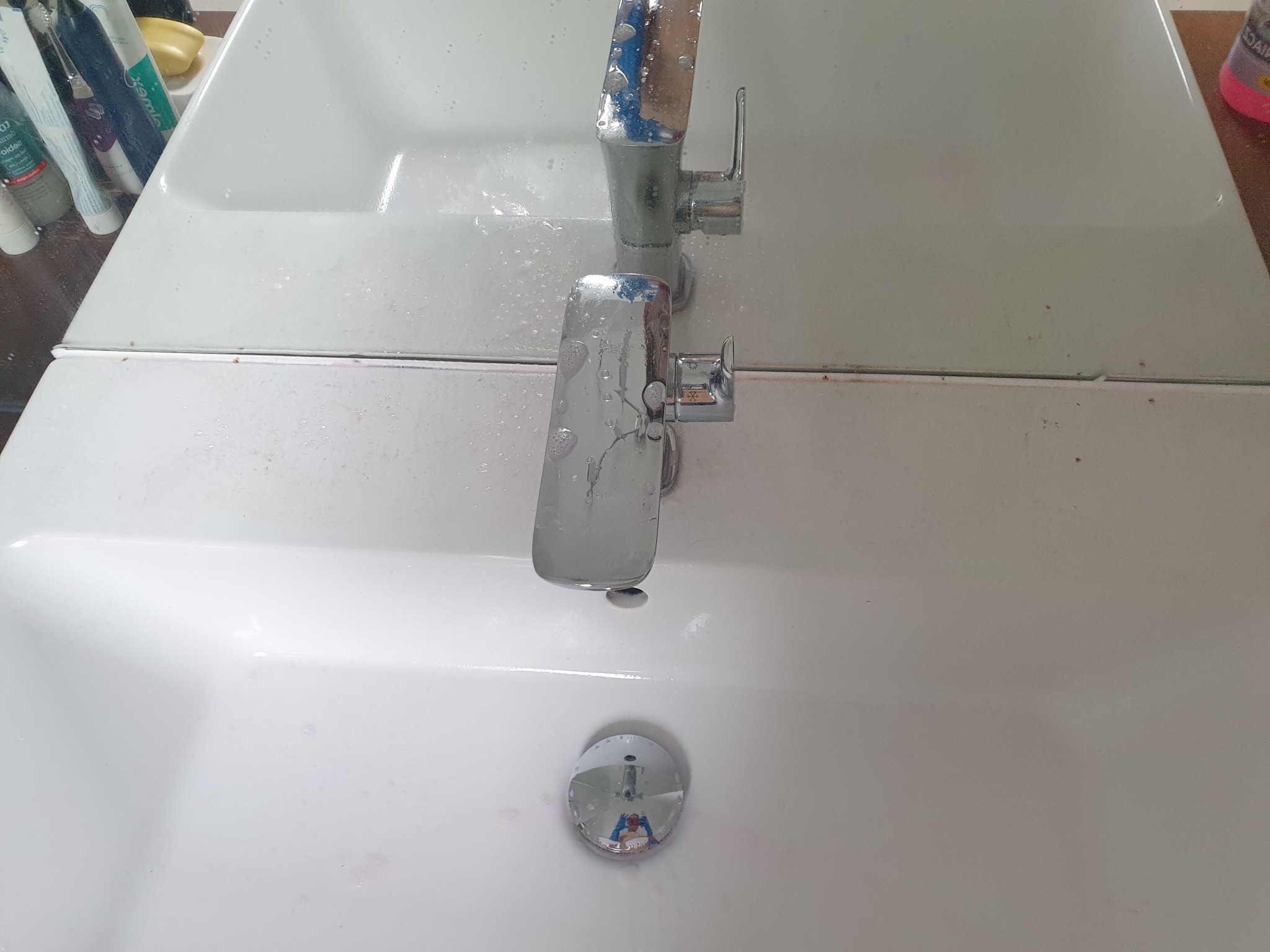 Biała umywalka z chromowaną baterią, na której widoczne są krople wody, a w odpływie odbija się osoba robiąca zdjęcie.