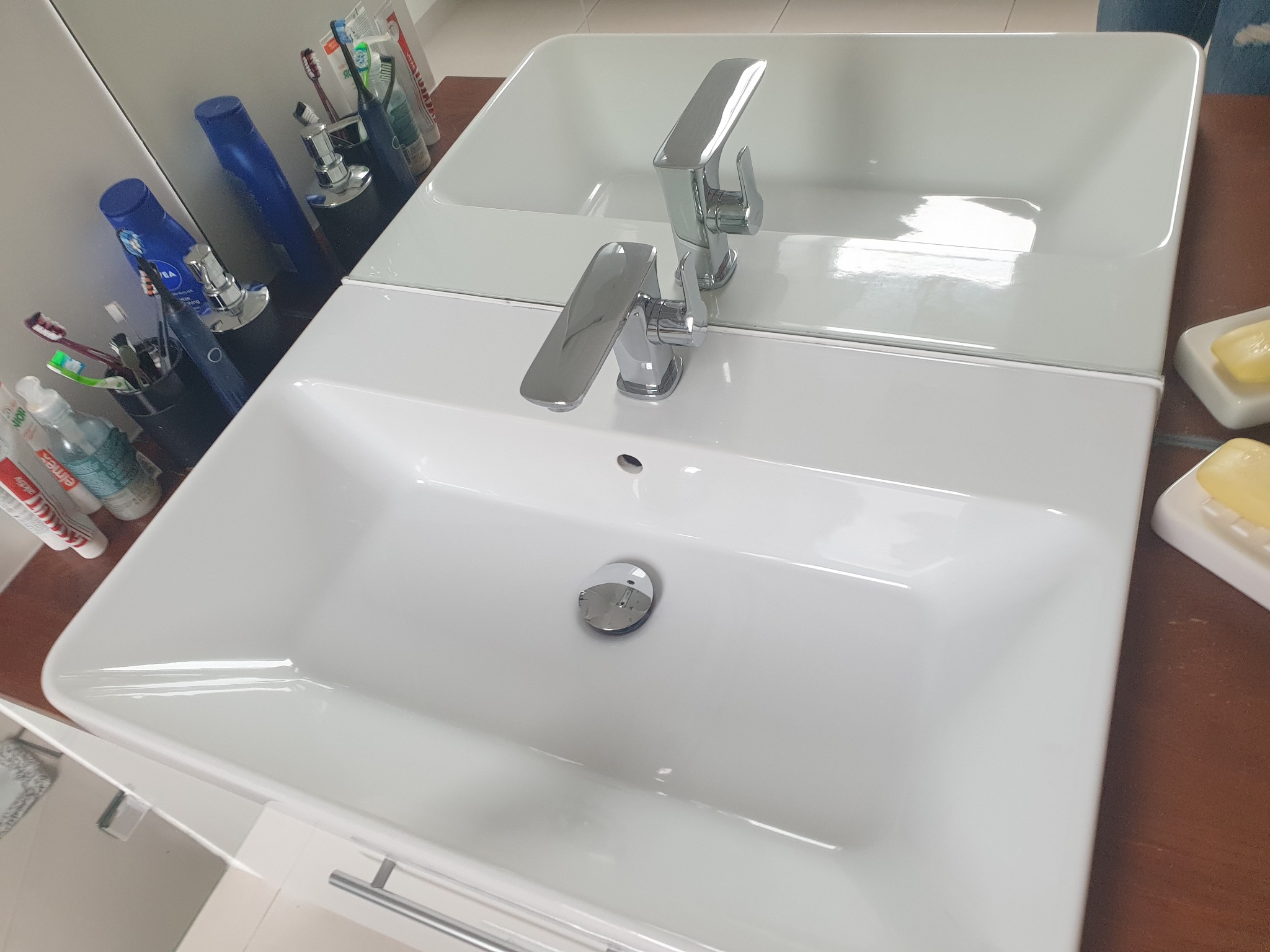 Biała, prostokątna umywalka z chromowaną baterią, odbijająca się w lustrze, z akcesoriami łazienkowymi w tle i mydłem w mydelniczce po prawej stronie.