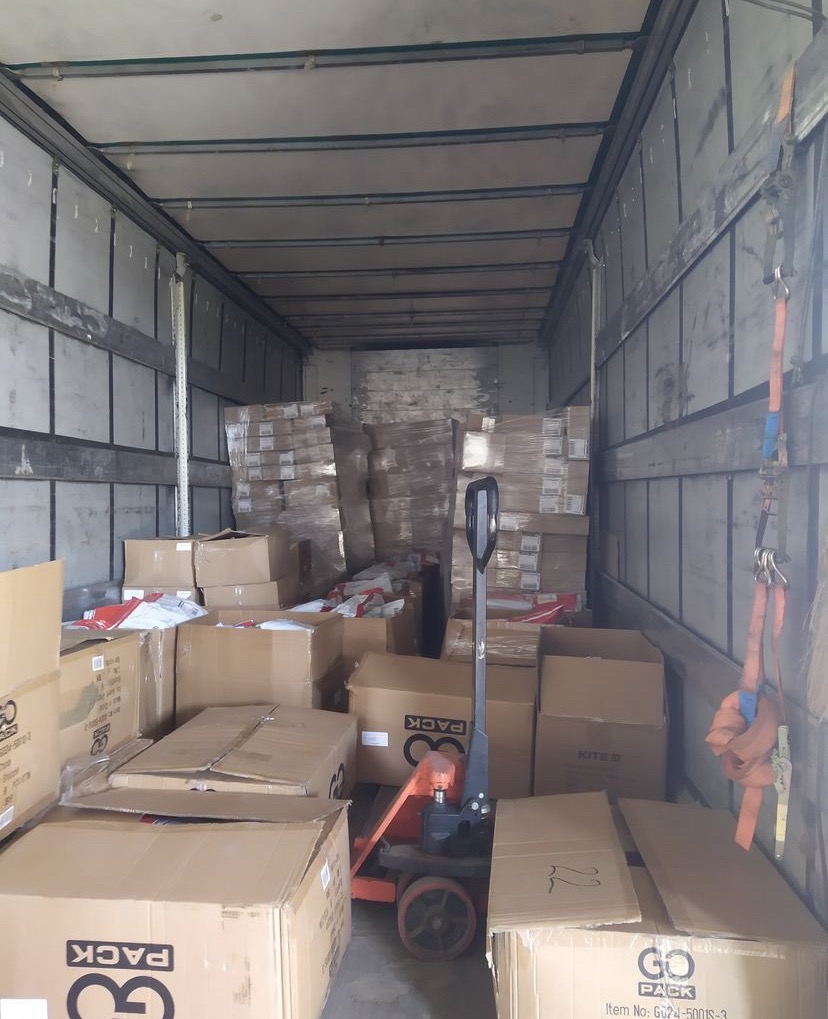 Wnętrze furgonetki wypełnionej kartonami i paletami z zapakowanymi towarami, z widocznym wózkiem paletowym pośrodku.