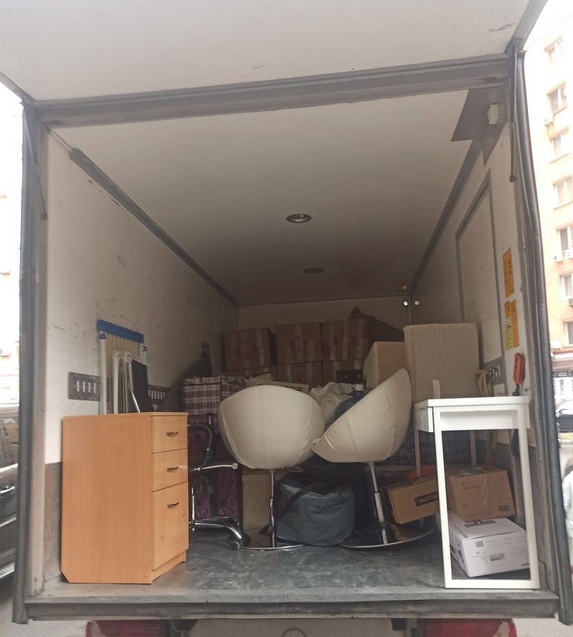 Wnętrze furgonetki załadowane meblami i kartonami podczas transportu, widoczne krzesła, biurko i komoda.