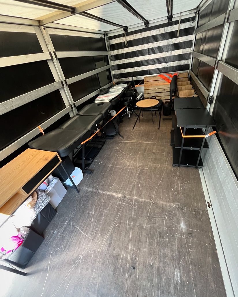 Wnętrze furgonetki z meblami i sprzętem, w tym kozetka do masażu, stolik, krzesła i regał zabezpieczone pomarańczowymi taśmami.