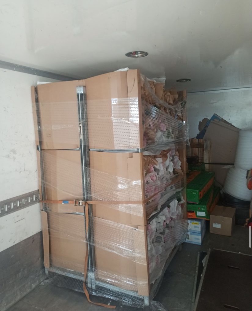 Kartonowe pudła zabezpieczone pasami i folią stretch na metalowym wózku wewnątrz przestrzeni ładunkowej samochodu dostawczego podczas transportu.