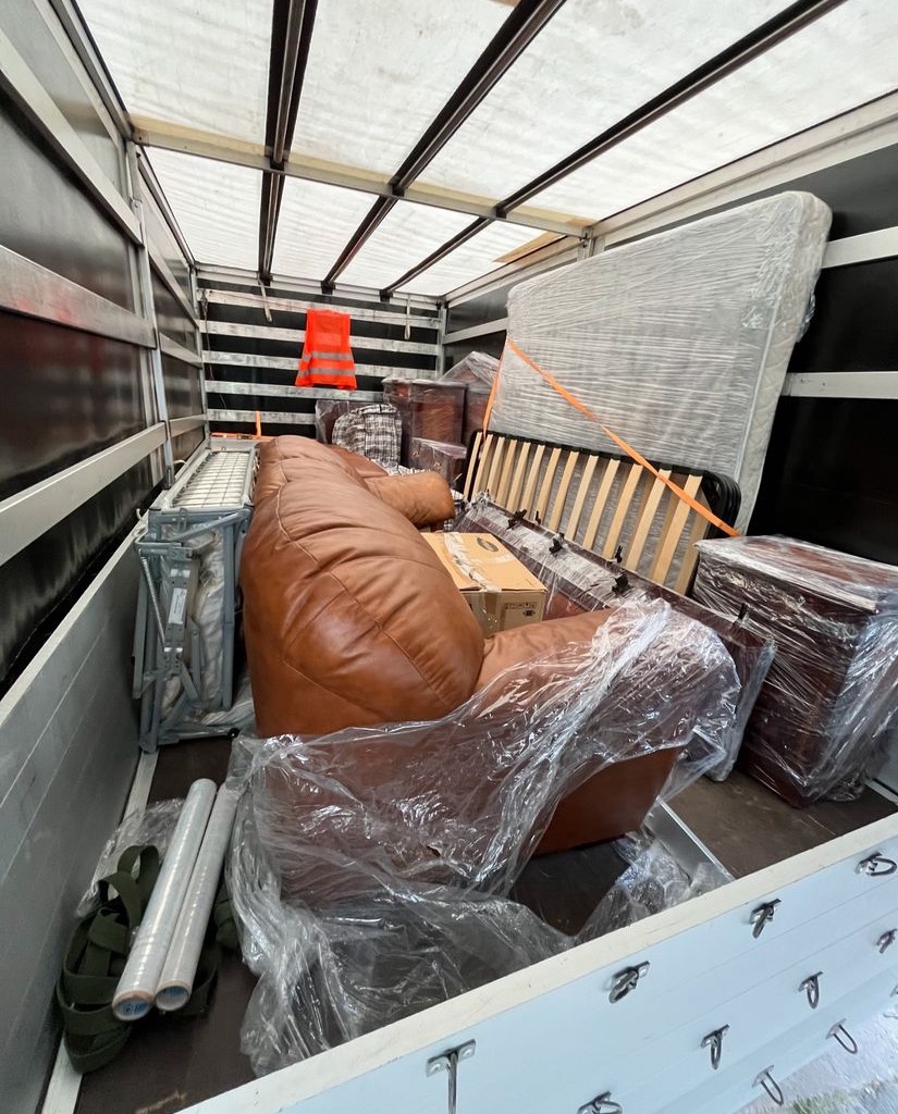 Wnętrze furgonetki załadowanej meblami, w tym brązowy, skórzany fotel typu worek, materac owinięty folią, rama łóżka i inne przedmioty zabezpieczone na czas transportu, a na ścianie kamizelka...