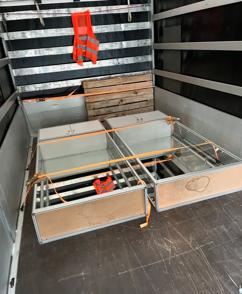 Wnętrze furgonetki z zabezpieczonymi dwoma metalowymi szafkami z szufladami, spiętymi pomarańczowymi pasami transportowymi, na podłodze widoczne ślady użytkowania, na ścianie wisi pomarańczowa...