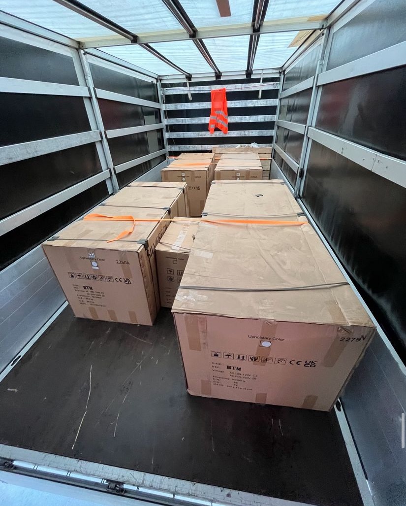 Wnętrze ciężarówki z kartonami zabezpieczonymi pomarańczowymi pasami transportowymi; na ścianie wisi pomarańczowa kamizelka odblaskowa.