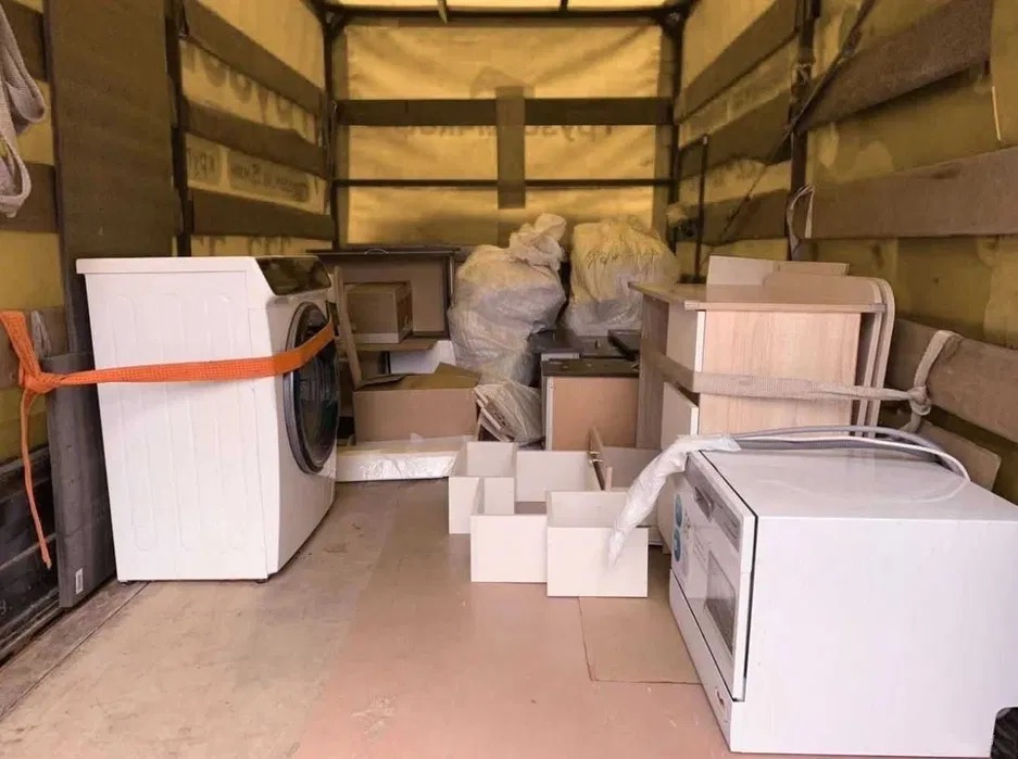 Wnętrze skrzyni ładunkowej wypełnionej meblami i sprzętem AGD, w tym pralka zabezpieczona pomarańczowym pasem transportowym, kartony i worki z dobytkiem podczas relokacji.