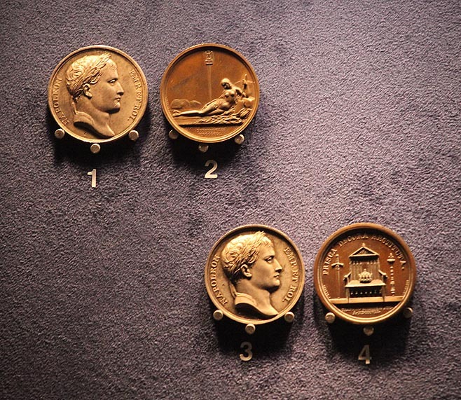 Cztery brązowe medale pamiątkowe z wizerunkiem Napoleona i alegorycznymi scenami, ponumerowane od 1 do 4, prezentowane na ciemnym, teksturowanym tle.