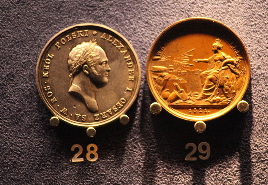 Dwie okrągłe, metalowe medale z wizerunkami i napisami, umieszczone na ciemnym, teksturowanym tle, ponumerowane 28 i 29.