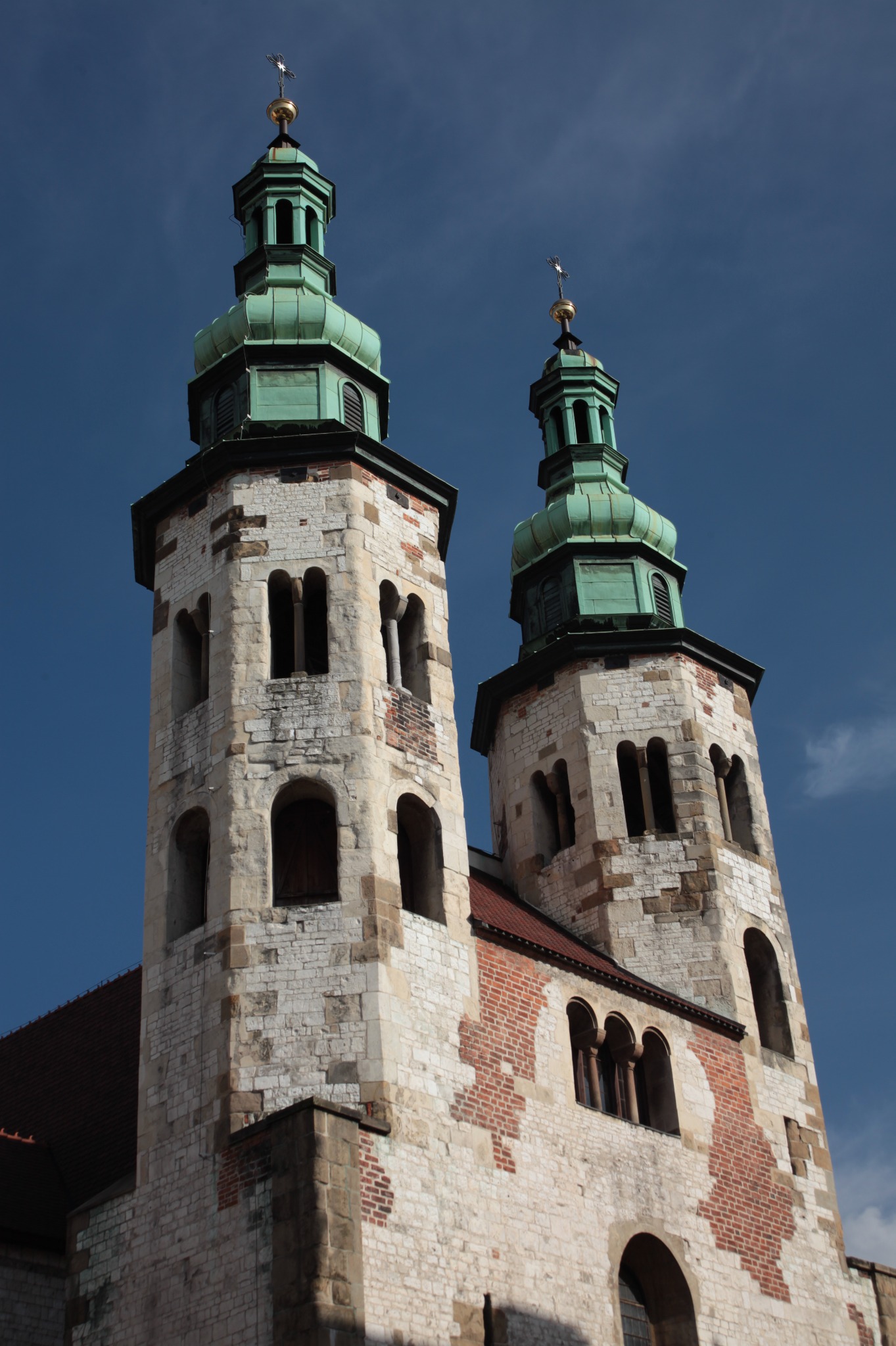 Dwie wieże kościoła z jasnego kamienia i cegły, zwieńczone zielonymi kopułami na tle błękitnego nieba.
