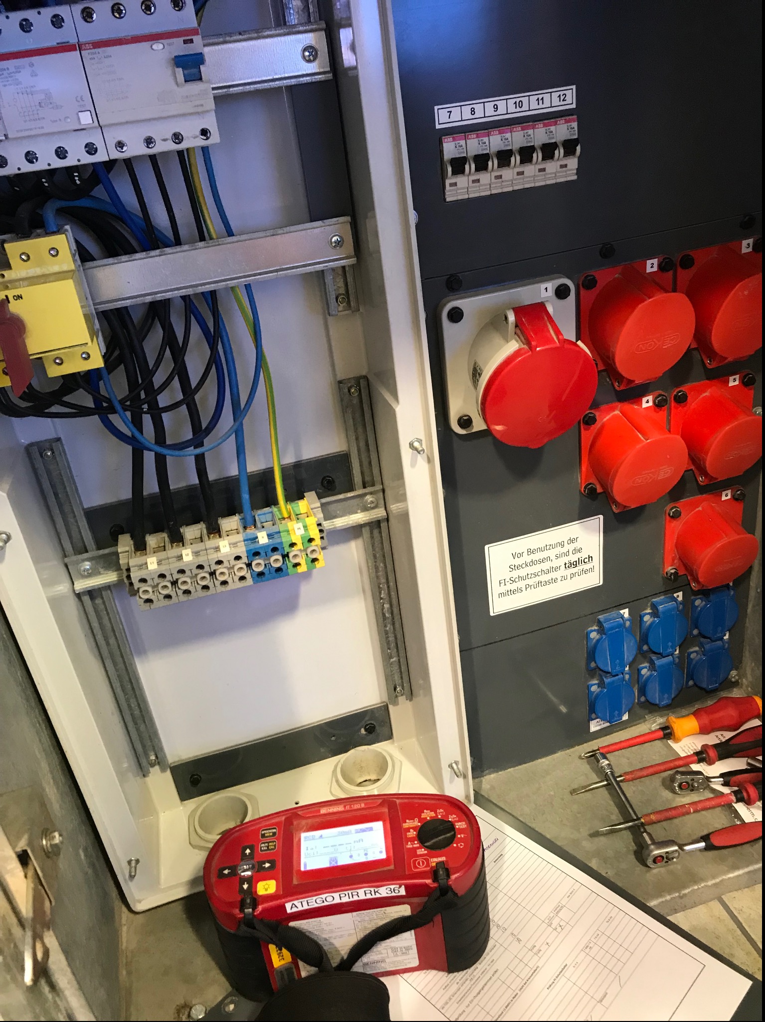 Otwarta skrzynka rozdzielcza z widocznymi przewodami, bezpiecznikami i złączkami, obok miernik ATEGO PIR RK 36 oraz narzędzia i wypełniony formularz pomiarowy.