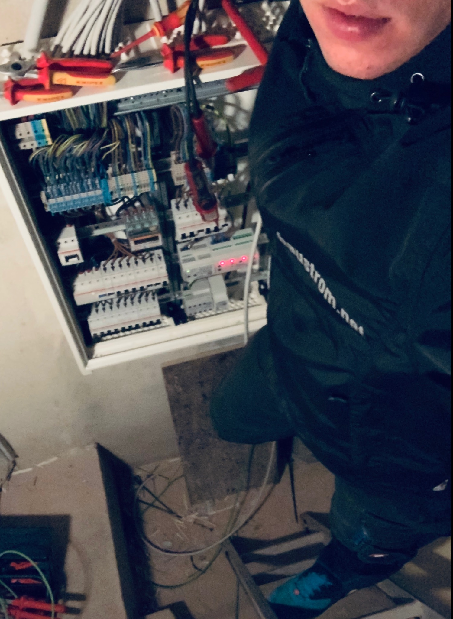 Elektryk w Oławie podczas prac w rozdzielnicy elektrycznej; widoczne kable, bezpieczniki, narzędzia i fragment twarzy elektryka w kurtce z logo.