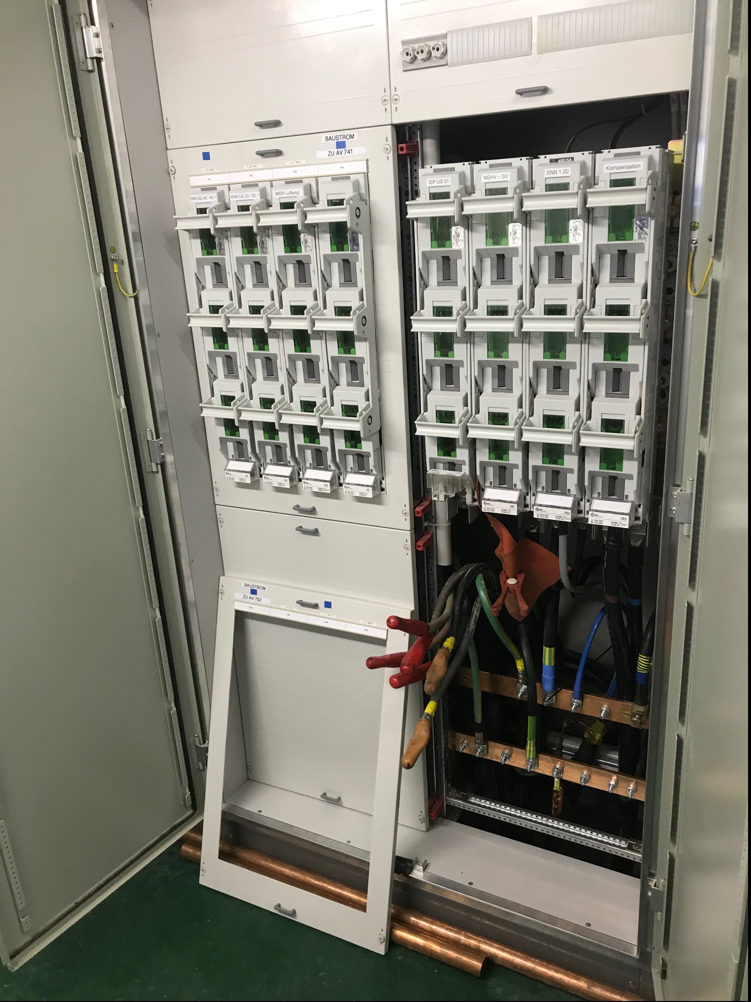 Otwarta szafa elektryczna z widocznymi bezpiecznikami, przewodami w różnych kolorach (czerwonym, niebieskim, zielonym i czarnym) oraz miedzianymi rurami na dole, z etykietami na poszczególnych...