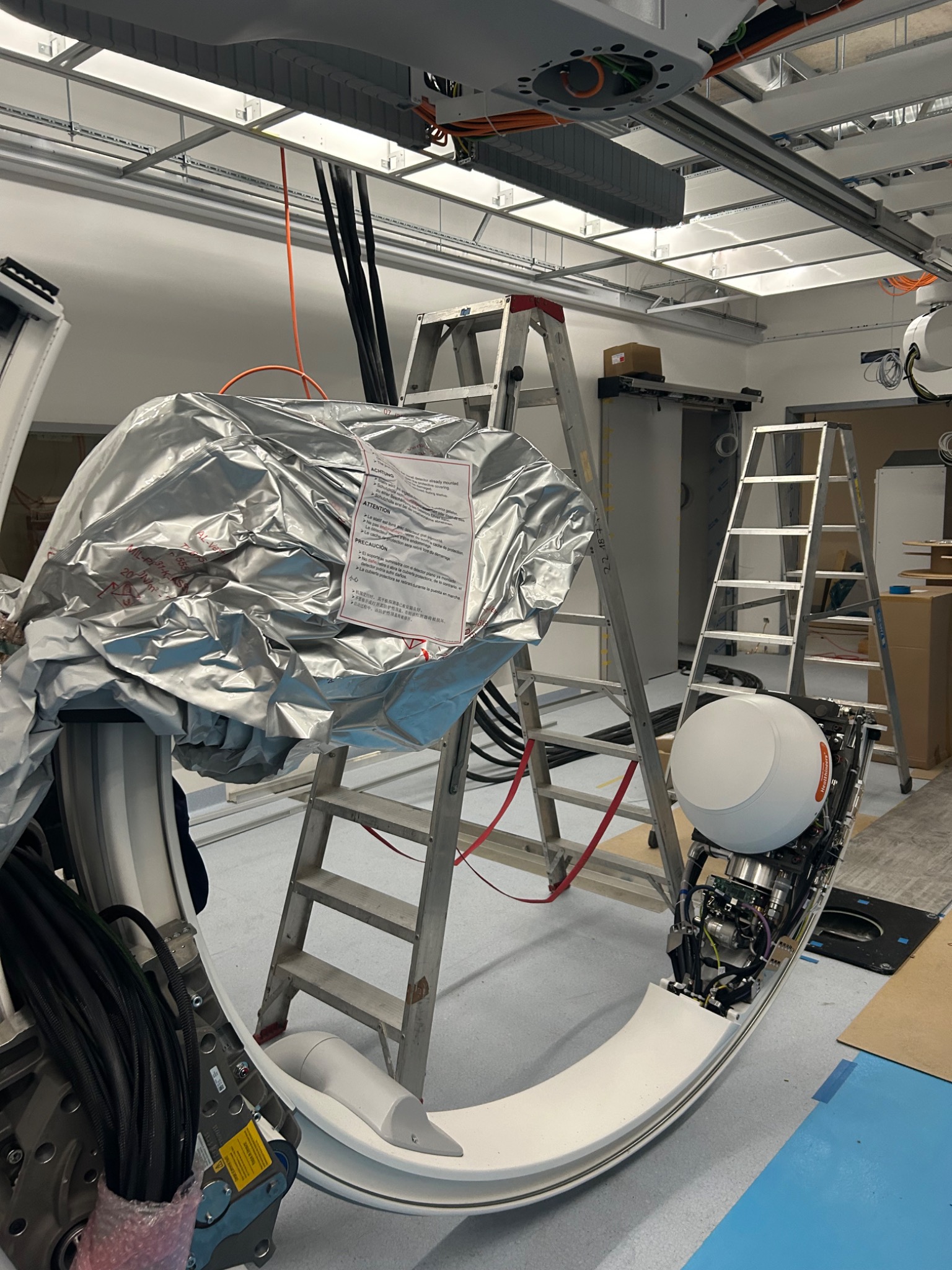 Wnętrze laboratorium z elementami instalacji elektrycznych, w tym drabiny, kable, osprzęt oświetleniowy oraz częściowo rozpakowany sprzęt medyczny z widocznymi przewodami i elektroniką.