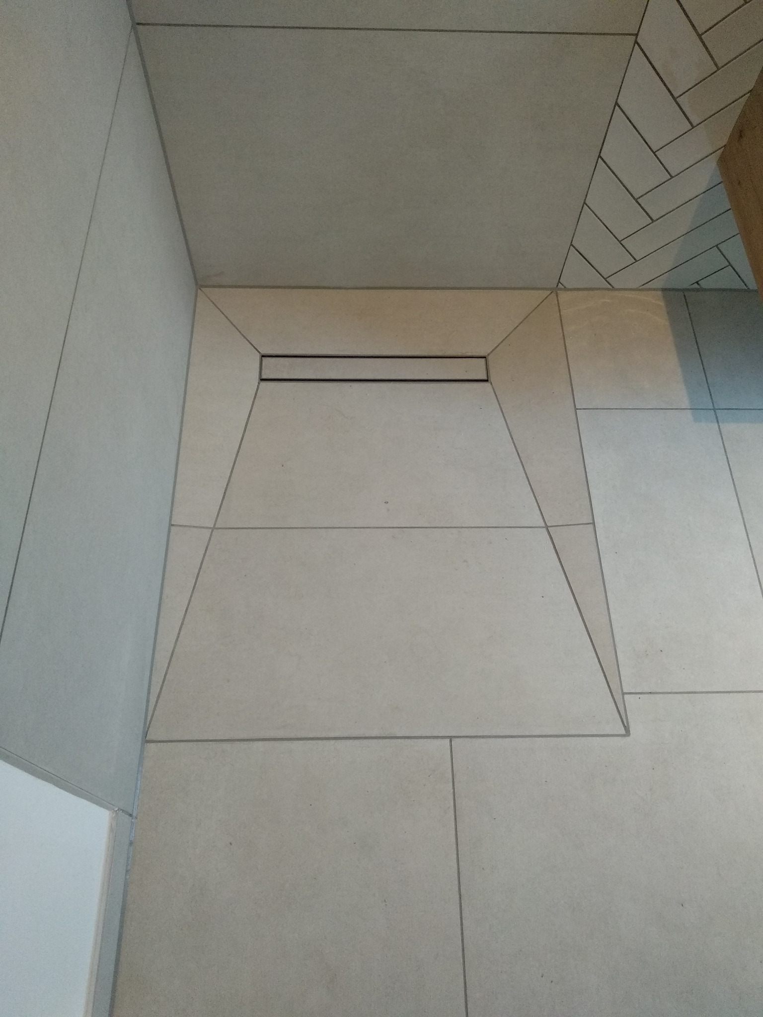 Beżowe płytki podłogowe ułożone wokół odpływu liniowego w kabinie prysznicowej w Wasilkowie, z fragmentem ściany wykończonej płytkami w jodełkę w tle.