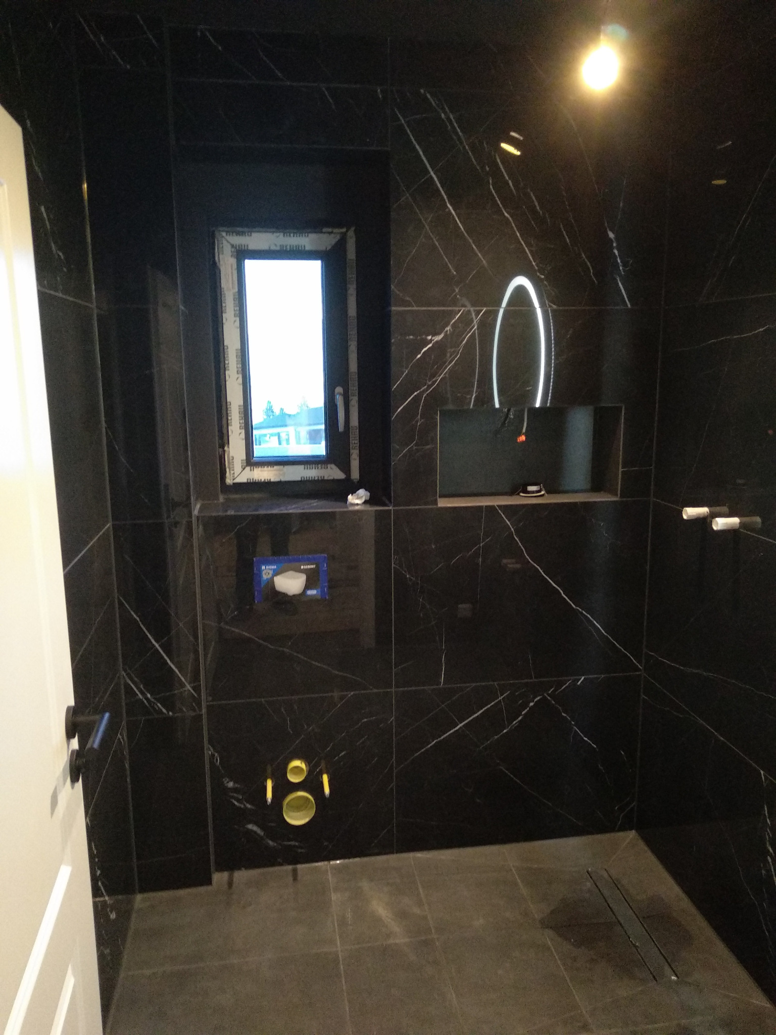 Nowoczesna łazienka wykończona czarnymi płytkami imitującymi marmur z białymi żyłami, z oknem, podświetlanym lustrem i odpływem liniowym w podłodze.