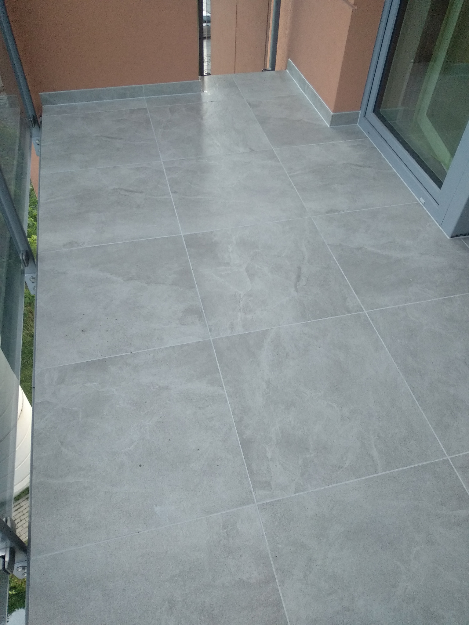 Jasnoszare płytki gresowe ułożone na balkonie, widoczne fugi i listwa przypodłogowa, szara rama okienna w tle.