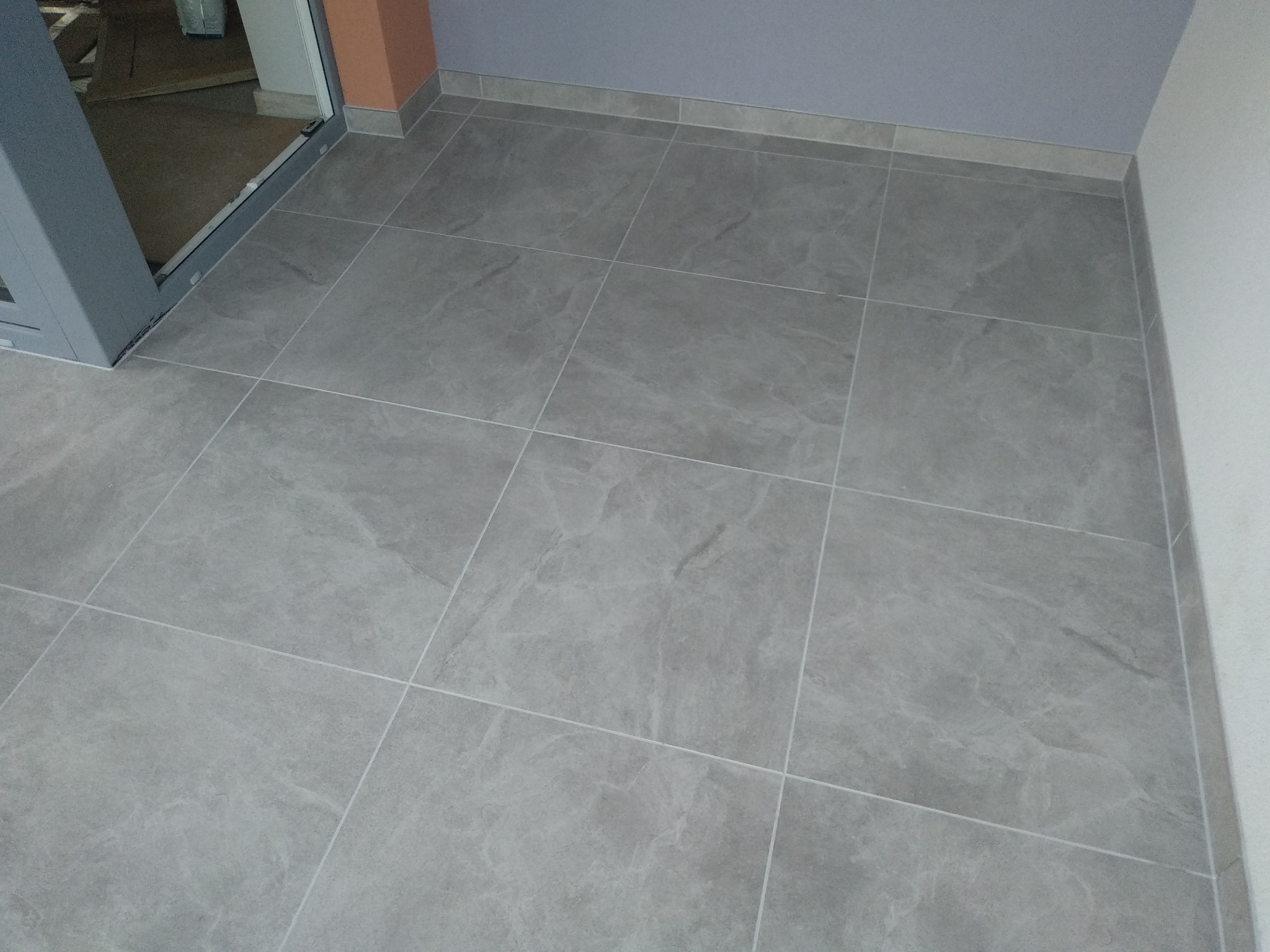 Ułożona szara podłoga z płytek imitujących kamień, z widocznymi fugami i listwami przypodłogowymi, ujęcie z góry w jasnym pomieszczeniu.