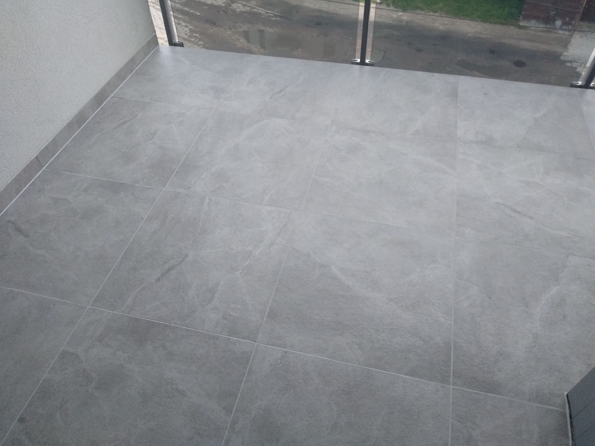 Szare płytki gresowe ułożone na balkonie, widok z góry, z widoczną szklaną balustradą w tle.