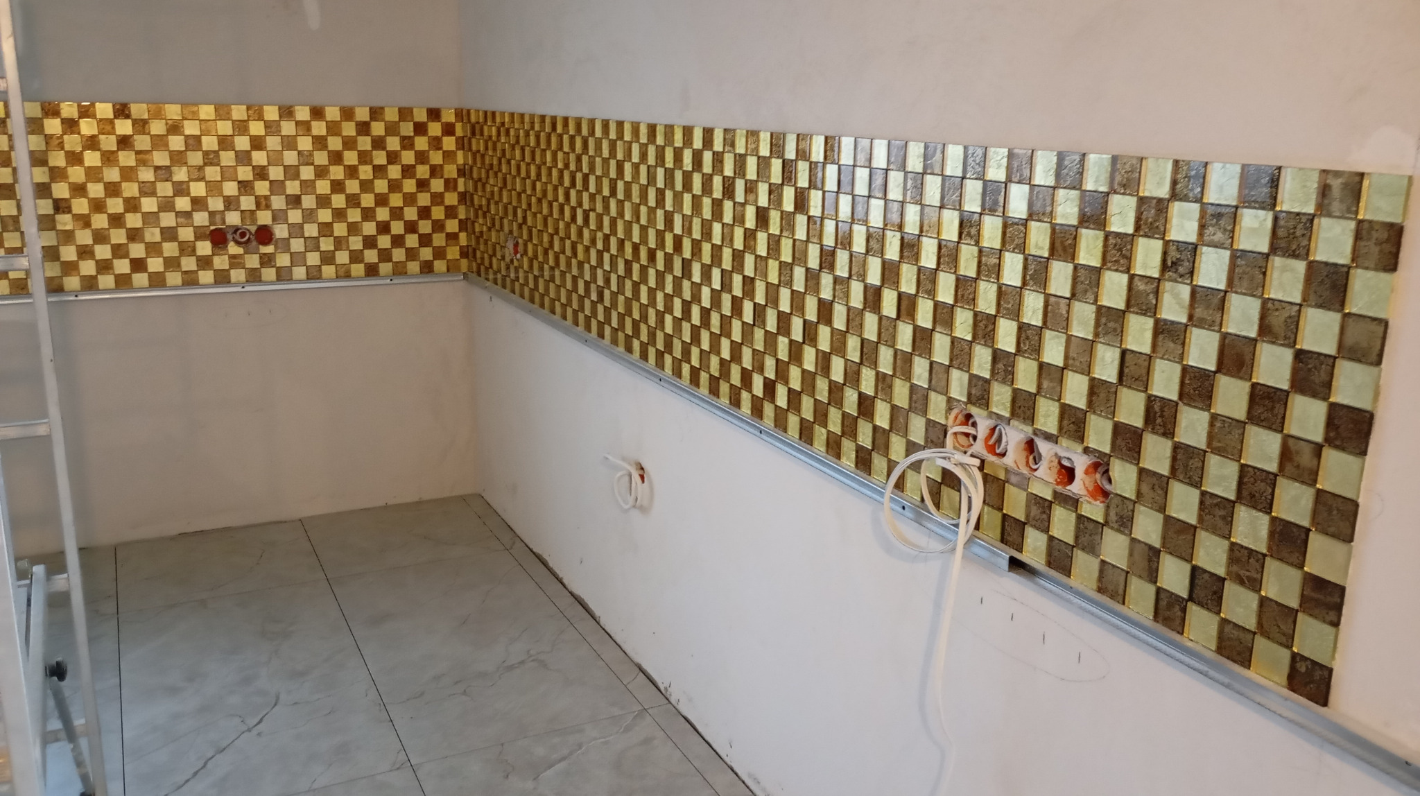 Ściana z mozaiki w szachownicę, brązowo-złota, częściowo ułożona w kuchni. Widoczne gniazdka elektryczne i drabina malarska. Podłoga wyłożona płytkami.