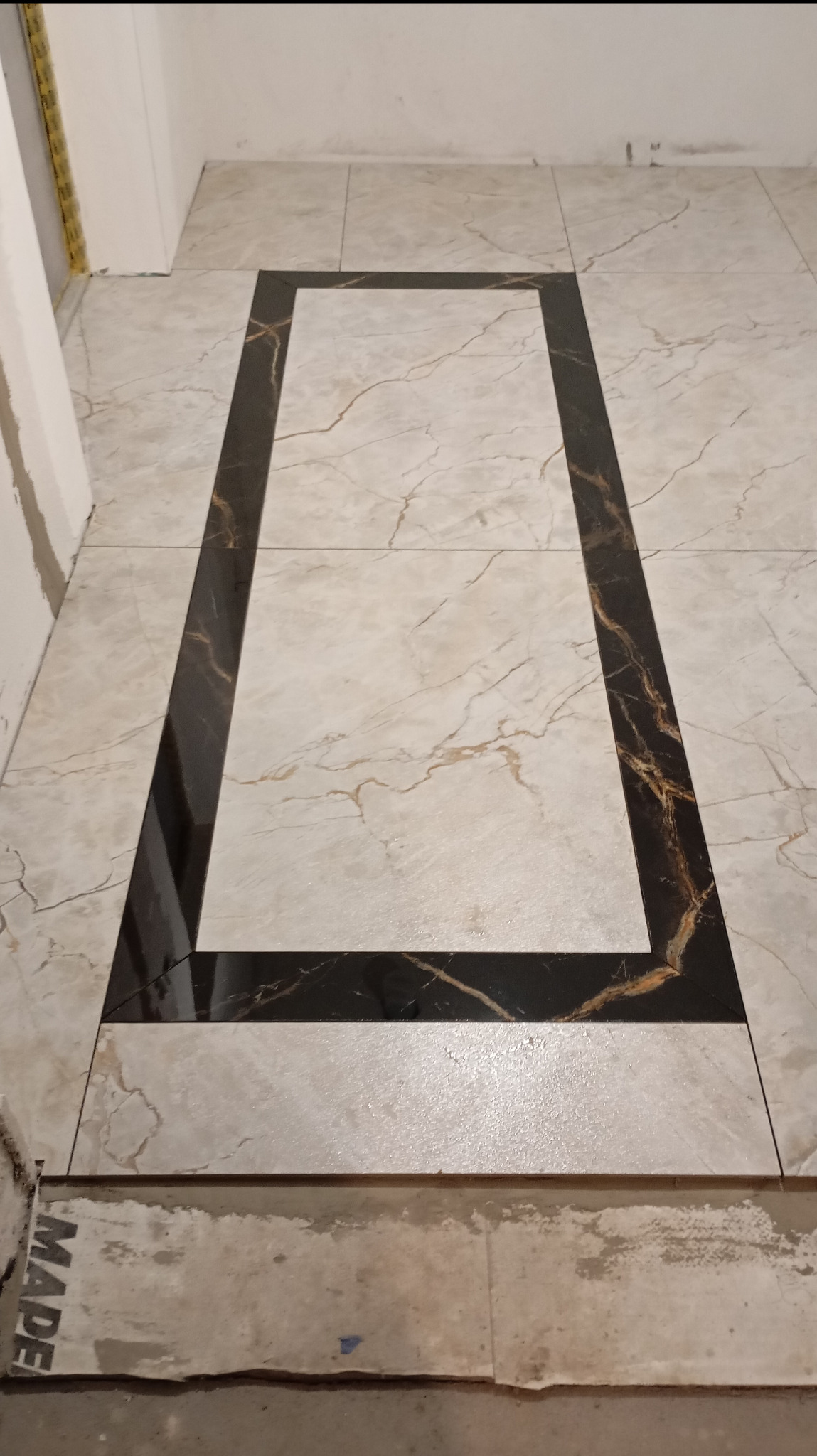 Ułożona podłoga z płytek w geometryczny wzór, z czarną obwódką i jasnym, marmurkowym wypełnieniem. Widoczne fugi i fragment progu.