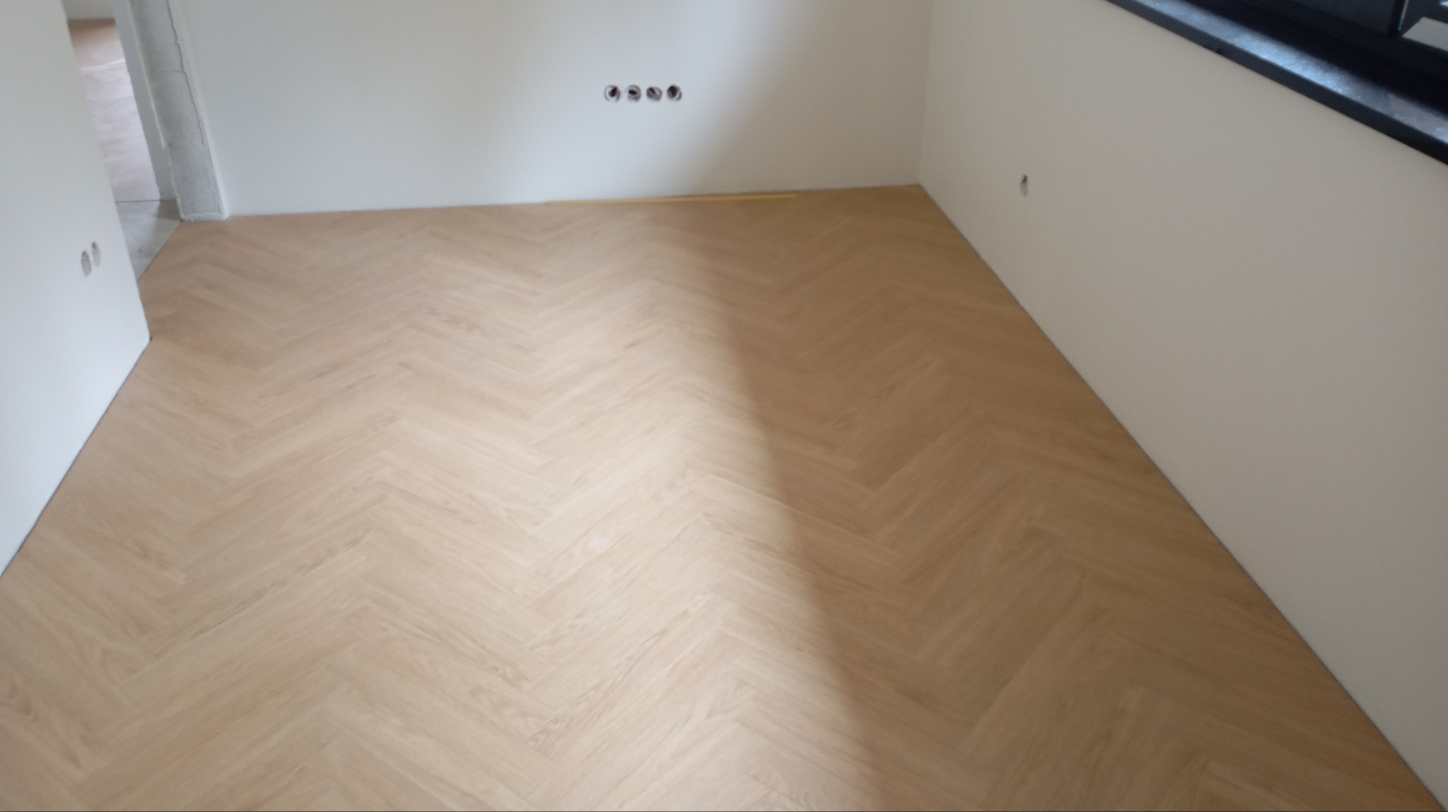 Jasna podłoga ułożona w jodełkę francuską w świeżo wykończonym pokoju z białymi ścianami i czarnym parapetem. Widoczne otwory na gniazdka elektryczne.