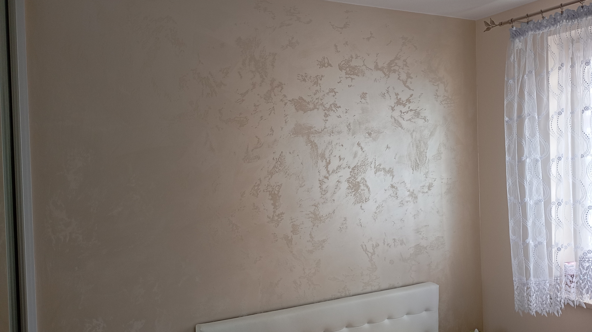 Ściana z dekoracyjną, beżową strukturą przypominającą wzór marmuru, widoczna w sypialni obok okna z firanką i fragmentu łóżka z białym zagłówkiem.