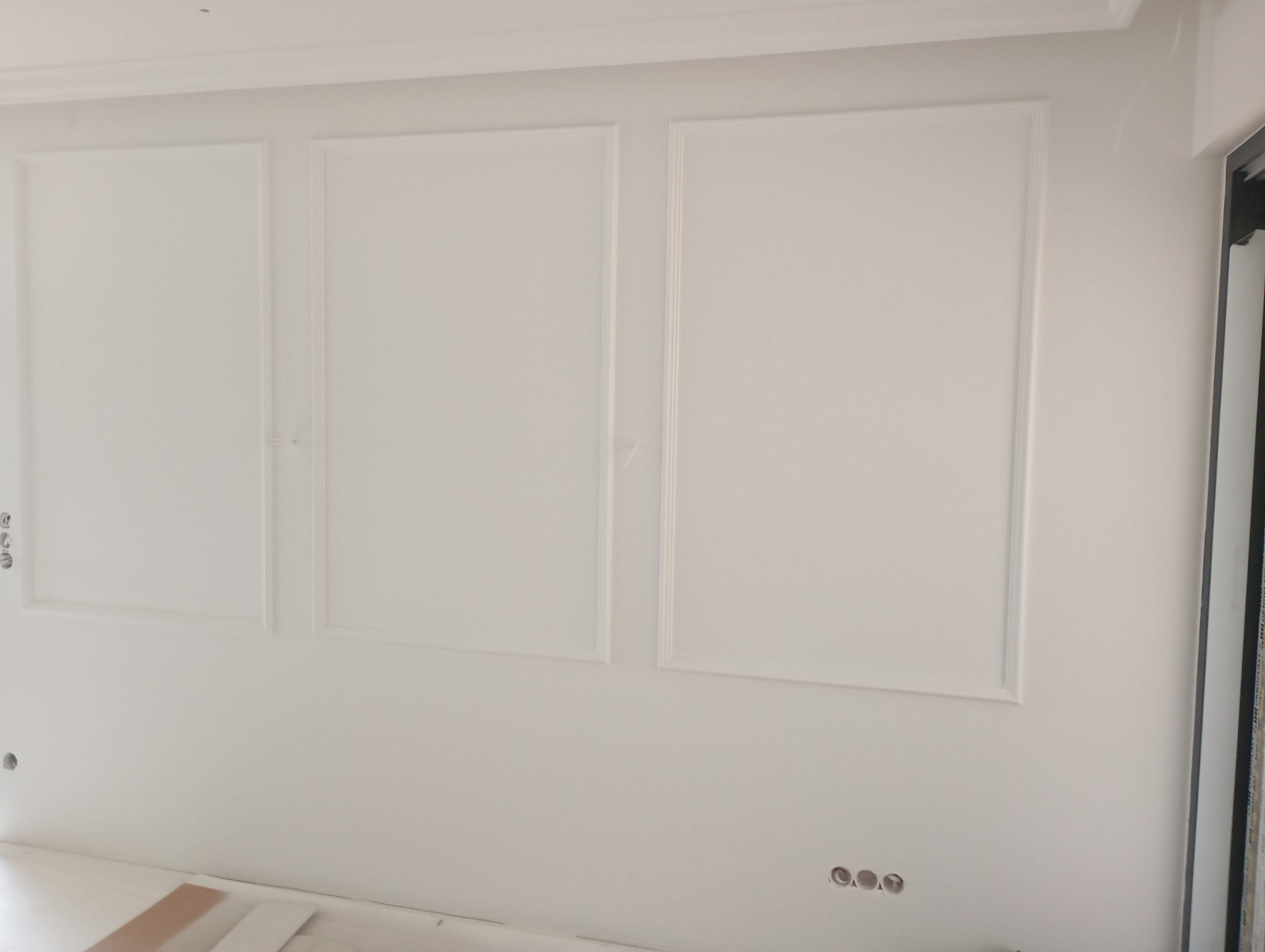 Biała ściana z trzema dekoracyjnymi panelami w ramkach, widoczne ślady po gniazdkach elektrycznych, listwa przypodłogowa w dolnej części kadru. Minimalistyczny design.