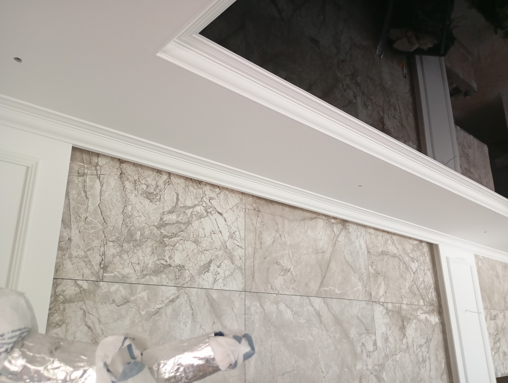 Wykończone wnętrze: marmurowe płytki na ścianie i biały sufit z ozdobnymi listwami. Widoczny fragment podwieszanego sufitu i częściowo zakryte folią elementy na dole.