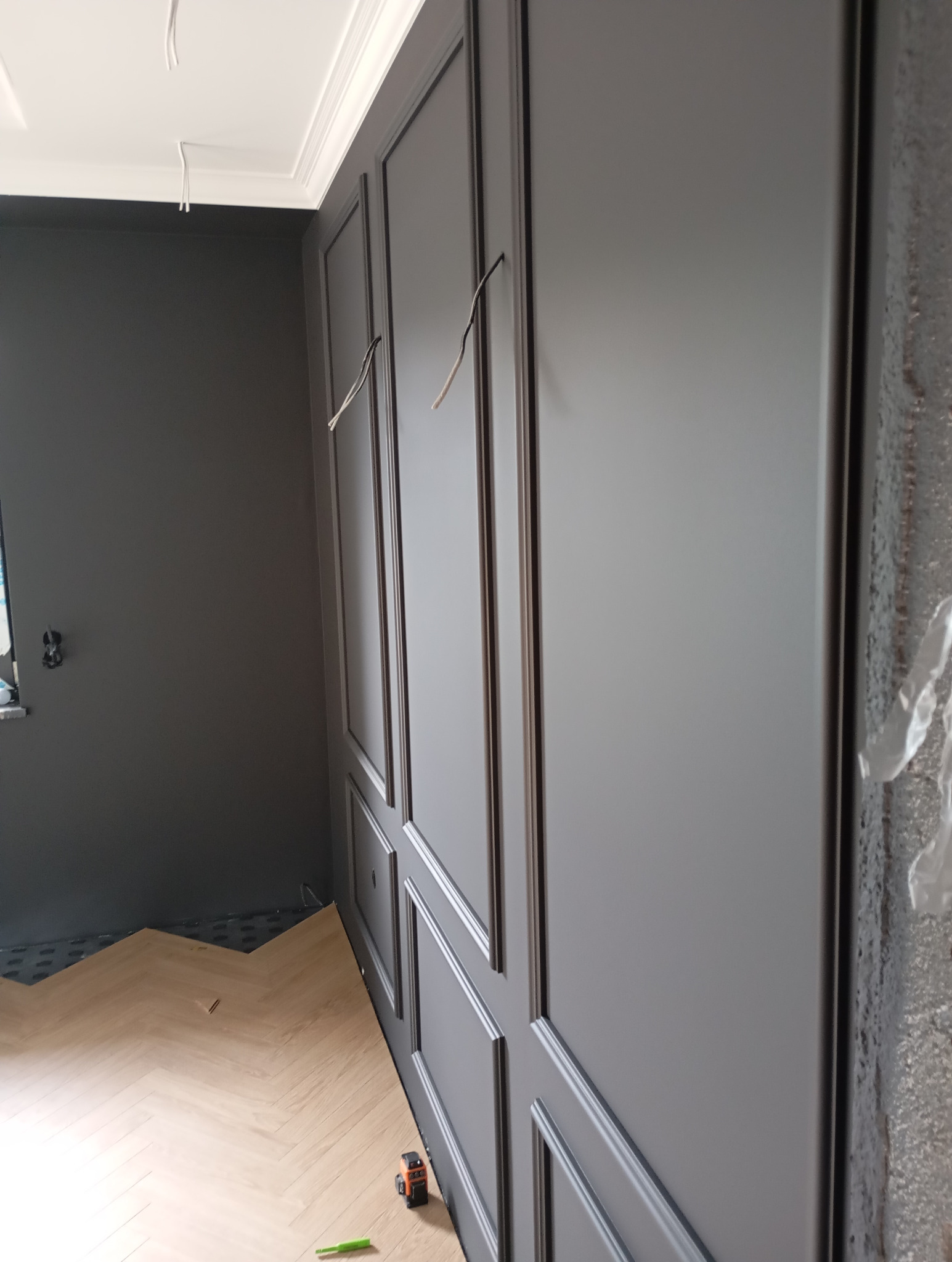 Elegancka ściana z panelami w odcieniach szarości, z widocznymi przewodami elektrycznymi i fragmentem ułożonej podłogi w jodełkę, sugerująca etap wykończenia wnętrza.