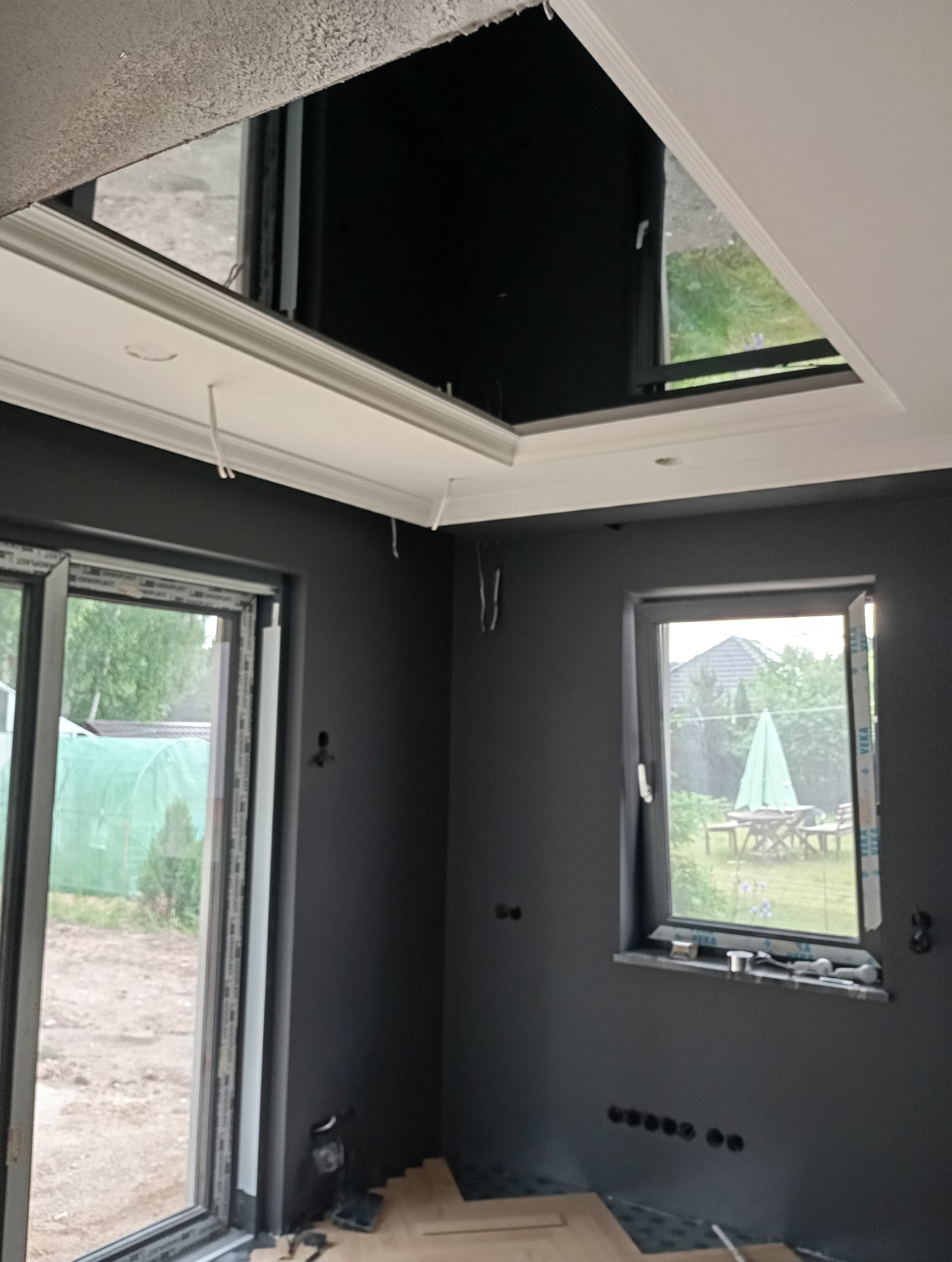 Wnętrze w trakcie wykańczania: szare ściany, częściowo ułożona podłoga, okno z widokiem na zewnątrz i otwór w suficie z ozdobną sztukaterią.