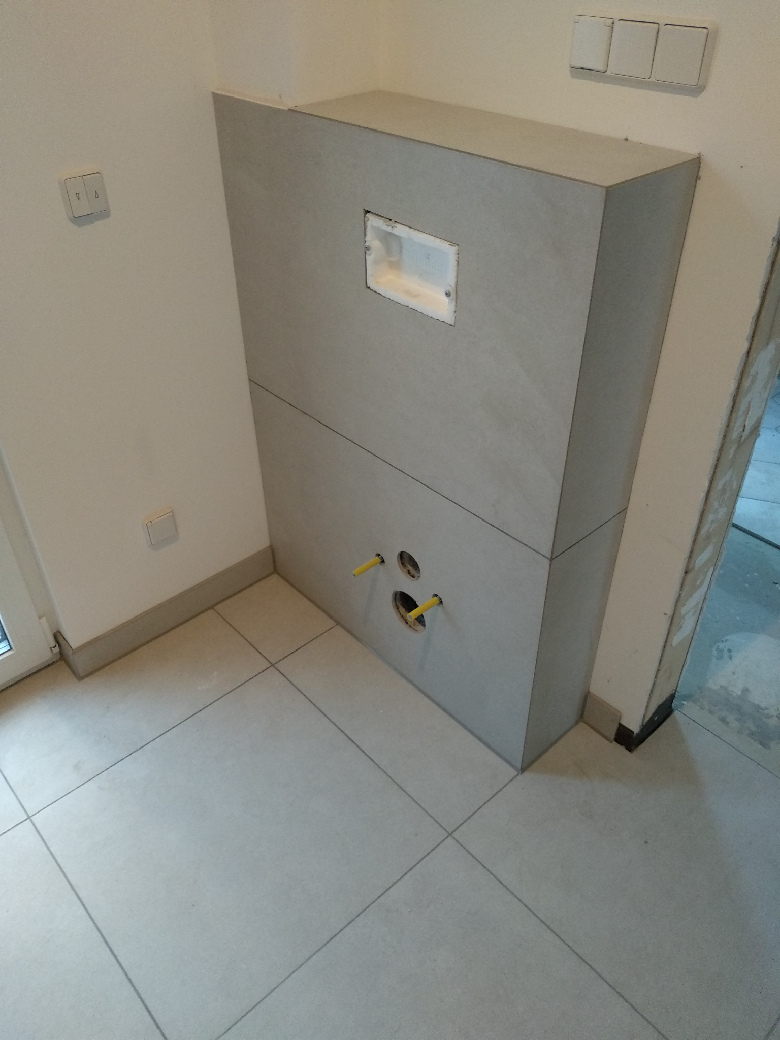 Zabudowa stelaża podtynkowego WC z płytkami 60x60, widoczne przyłącza wodne i otwór rewizyjny. Jasne płytki podłogowe i ściany. Element wykończenia łazienki.