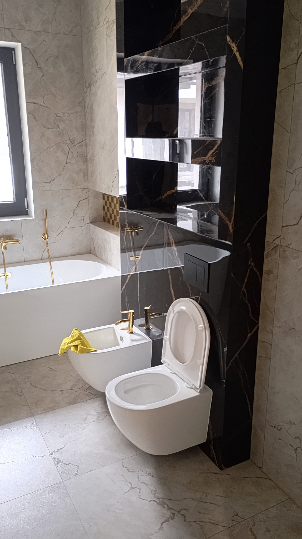 Elegancka łazienka z marmurowymi płytkami, złotą armaturą i wiszącą toaletą. Bidet z żółtym ręcznikiem. Nowoczesny design z czarnym akcentem półek.