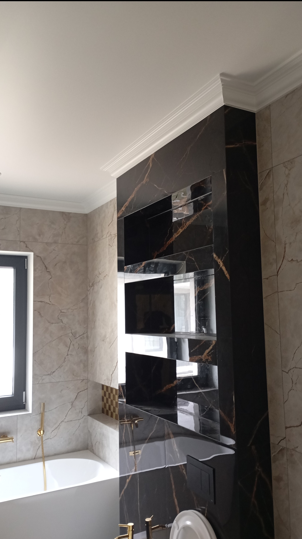 Elegancka łazienka z płytkami imitującymi marmur, czarna zabudowa z półkami i złotą armaturą, widok na wannę i okno. Nowoczesny wystrój z dbałością o detale.