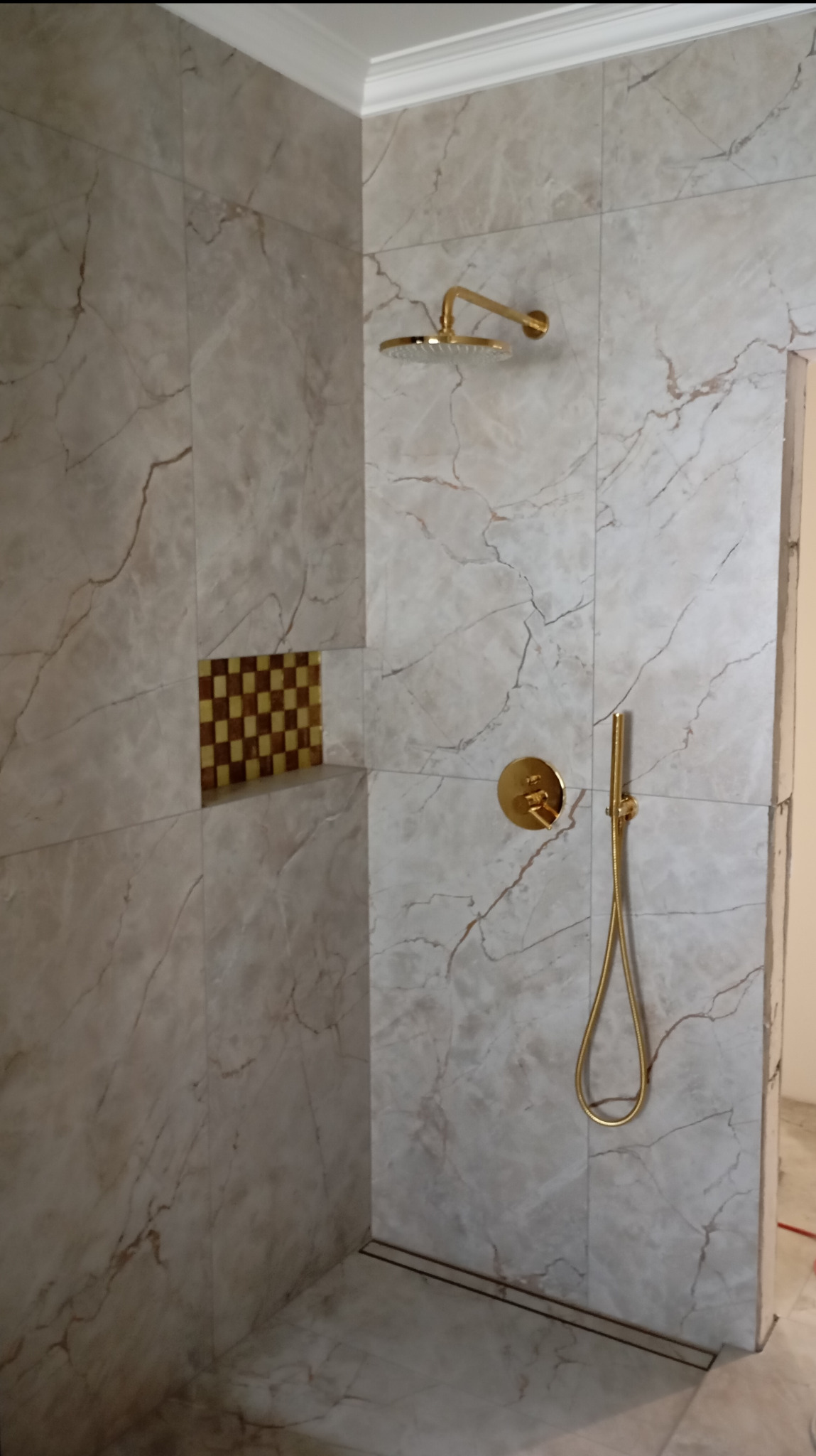 Wykończony prysznic z marmurowymi płytkami i złotą armaturą. W ścianie wnęka z mozaiką w szachownicę. Uwagę przykuwa odpływ liniowy w podłodze.