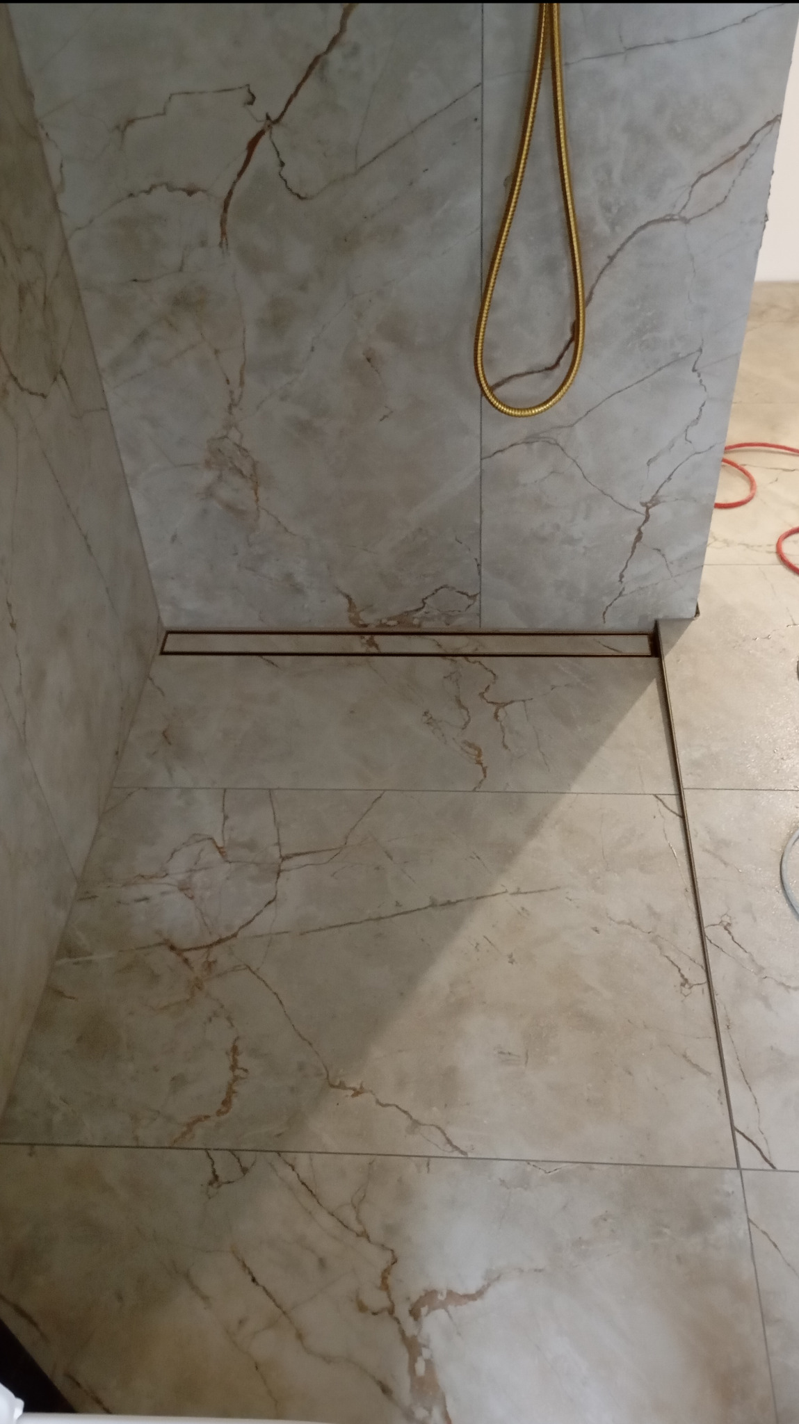 Ukończona łazienka z beżowymi płytkami imitującymi marmur i złotym odpływem liniowym. Widok na podłogę i fragment ściany.