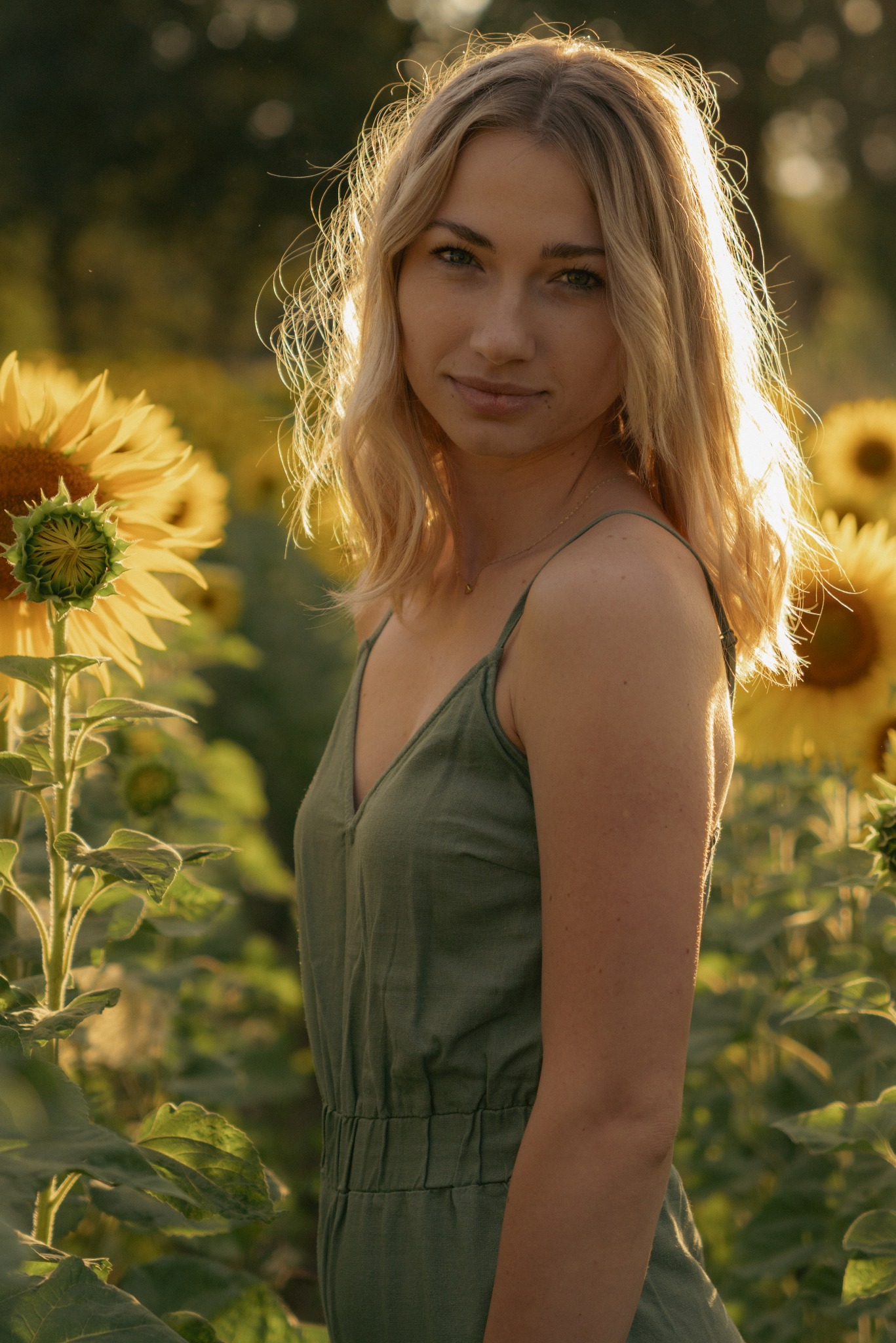 Portret kobiety w zielonym kombinezonie na tle pola słoneczników, oświetlona ciepłym, zachodzącym słońcem, z delikatnym uśmiechem i blond włosami opadającymi na ramiona.